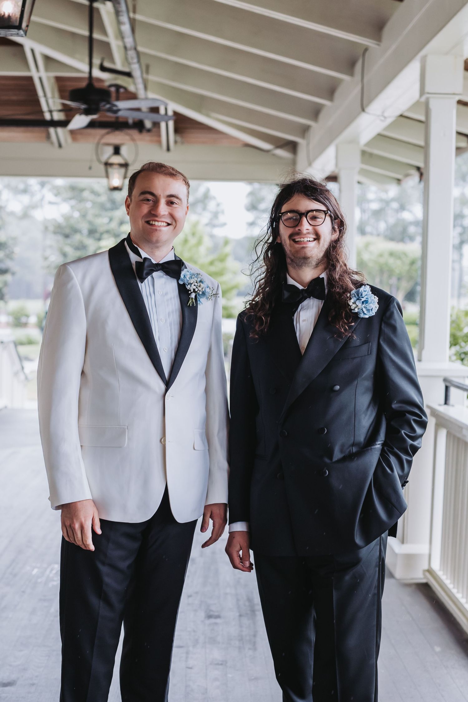 Two groomsmen in tuxedos with blue boutonnieres posing on covered porch at wedding.