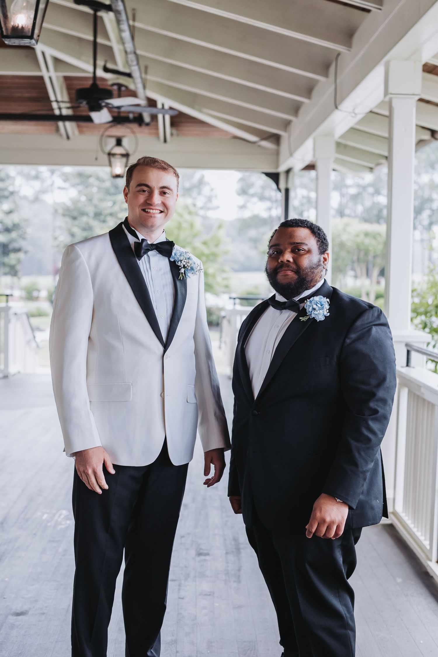 Groom in white tuxedo jacket poses with groomsman in black suit on wedding venue porch.