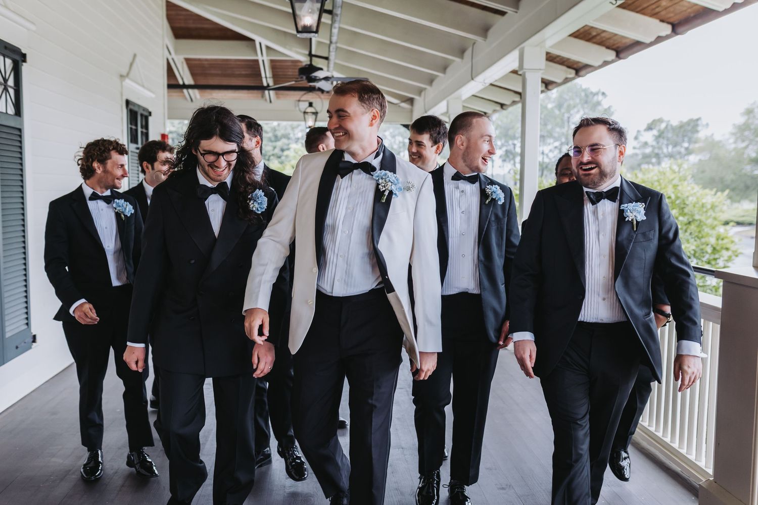 Groom in white jacket walks with groomsmen in black tuxedos along a covered porch.