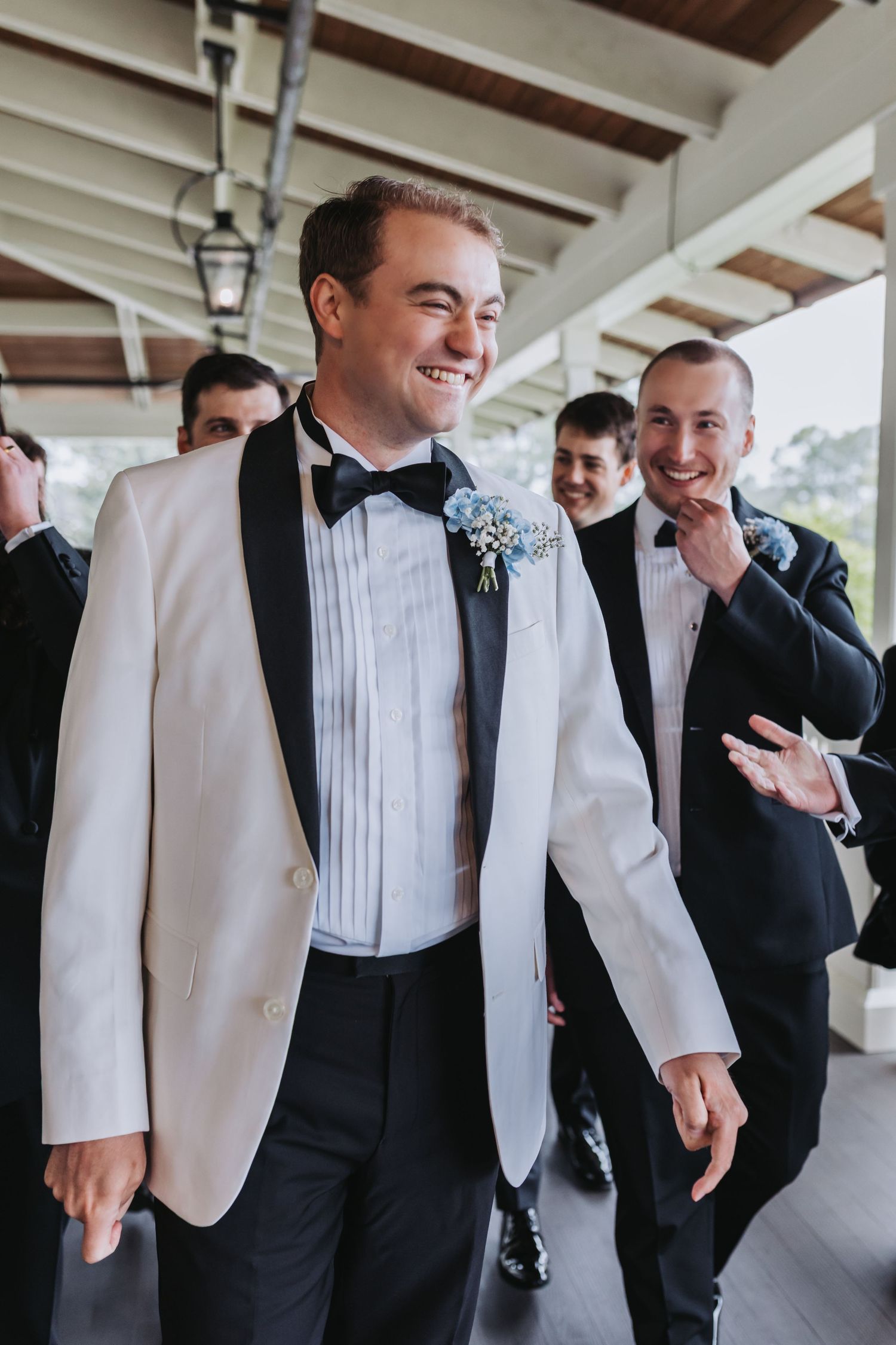 Groom in white tuxedo leads smiling groomsmen in black suits along covered venue walkway.