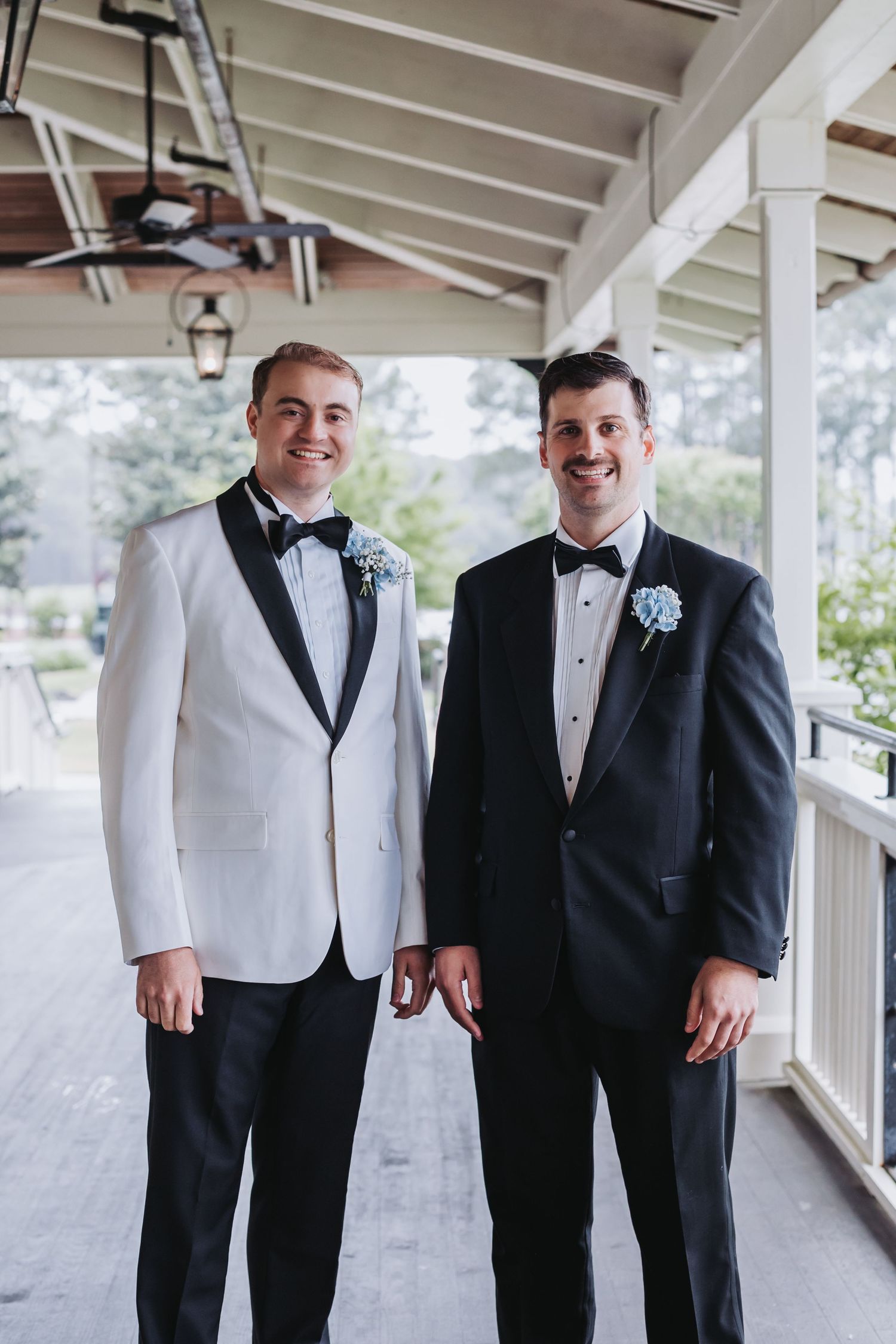 Groom in white tuxedo and best man in black tuxedo smile together on decorated outdoor porch.