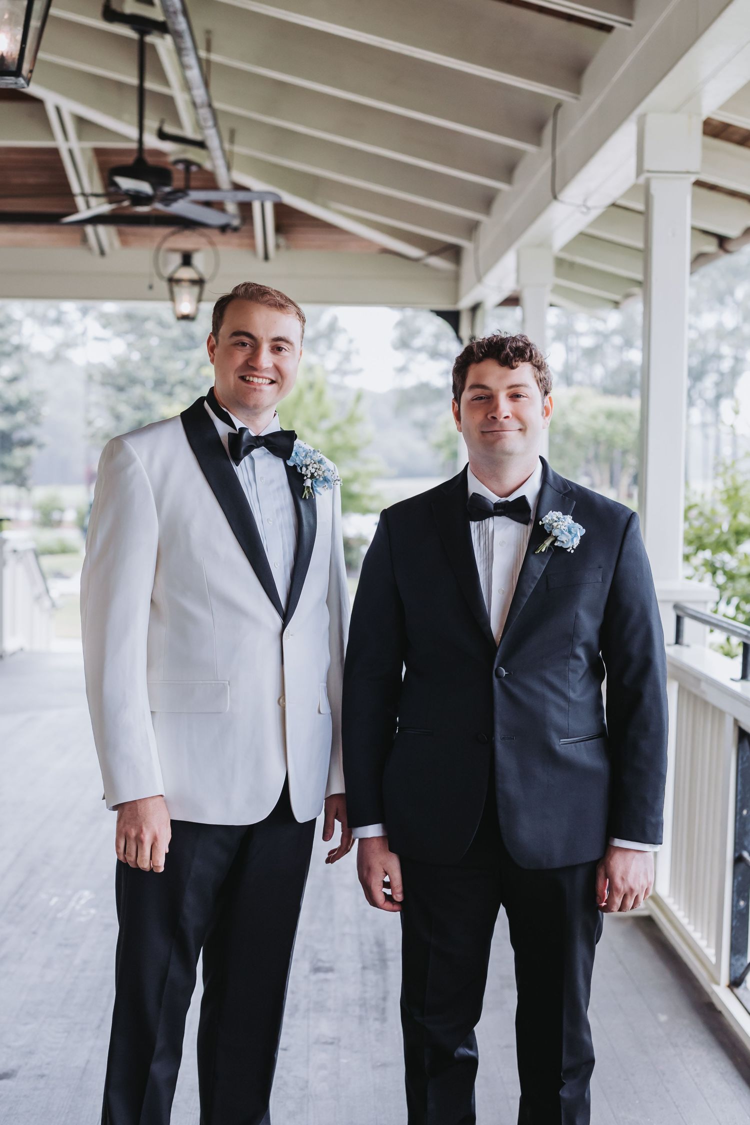 Two groomsmen in tuxedos with boutonnieres posing on a covered porch.
