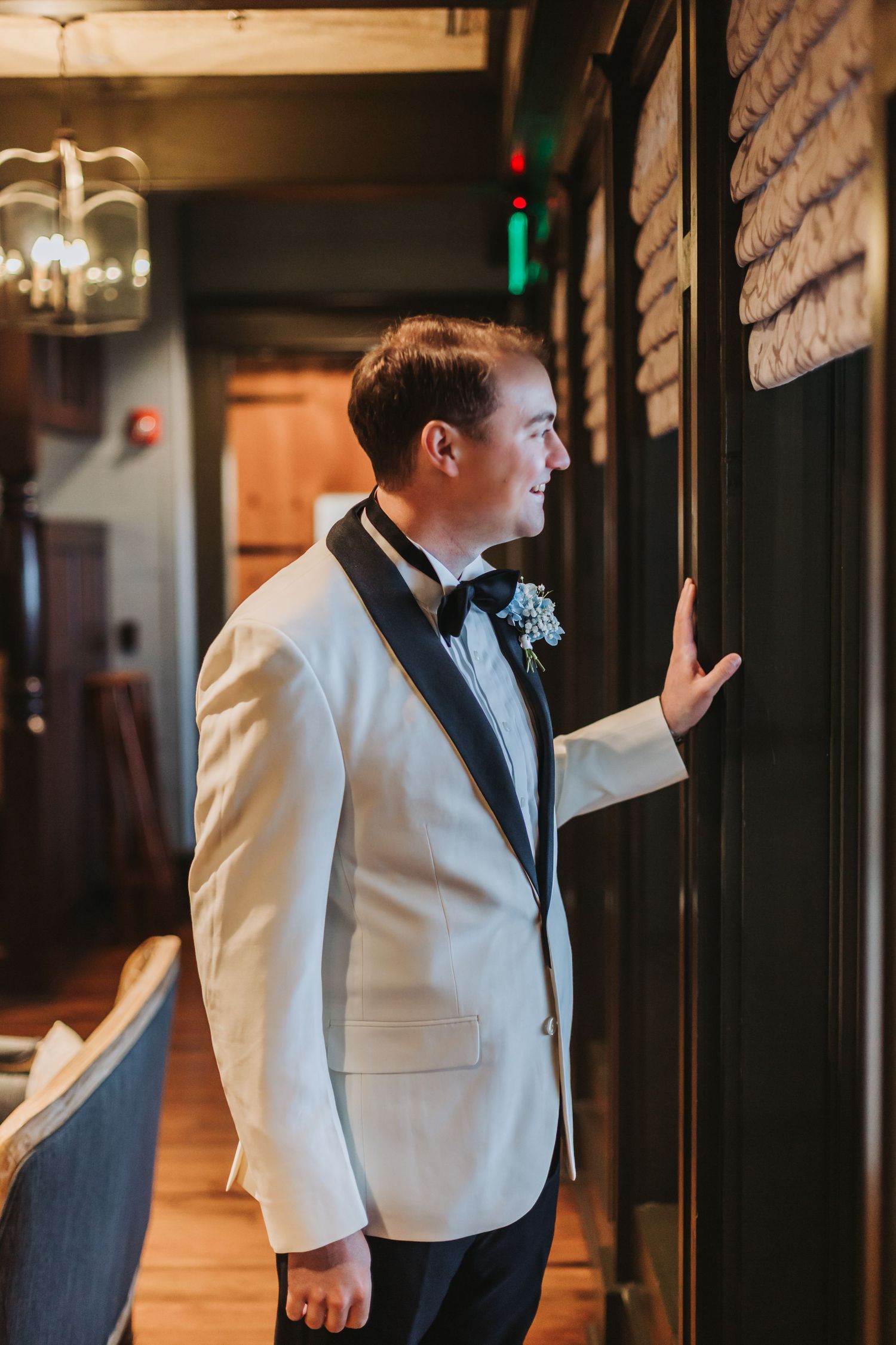 Groom in white dinner jacket with boutonniere standing by a dark wooden door.