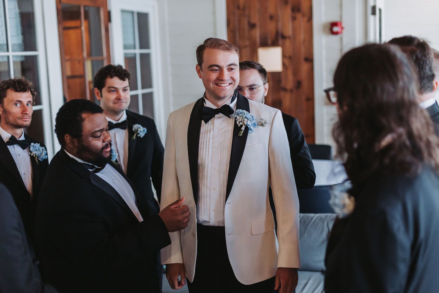 Groom in white tuxedo jacket smiles with groomsmen in black suits at wedding venue.