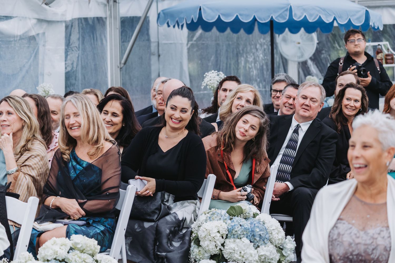 Wedding guests seated under a blue and white tent smile during an outdoor ceremony.