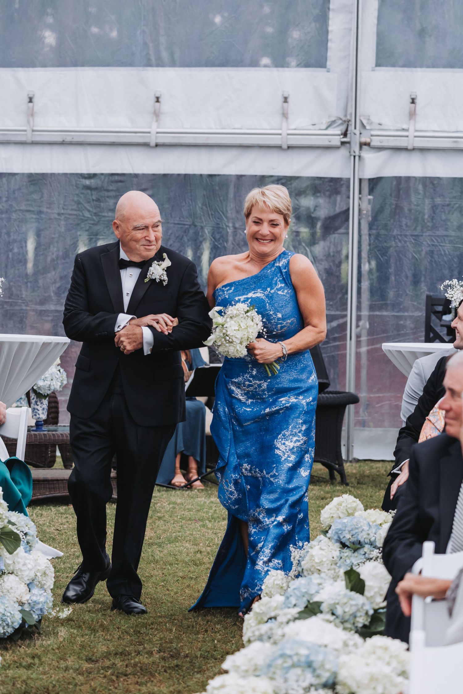 Couple walks down the aisle, woman in blue floral gown carrying white bouquet under a clear tent.