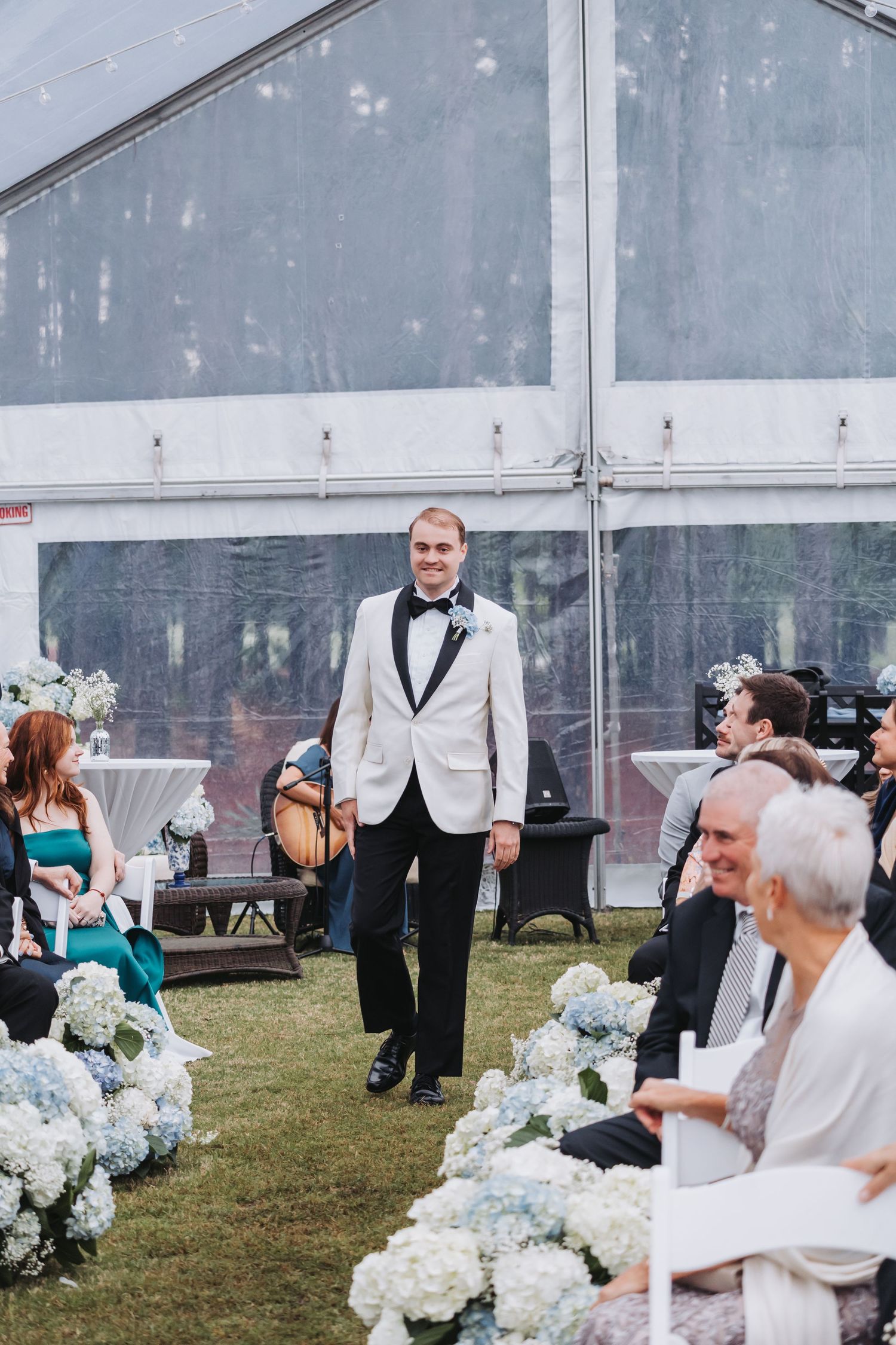 Smiling groom in white tuxedo jacket walks down flower-lined aisle at outdoor wedding ceremony under clear tent.
