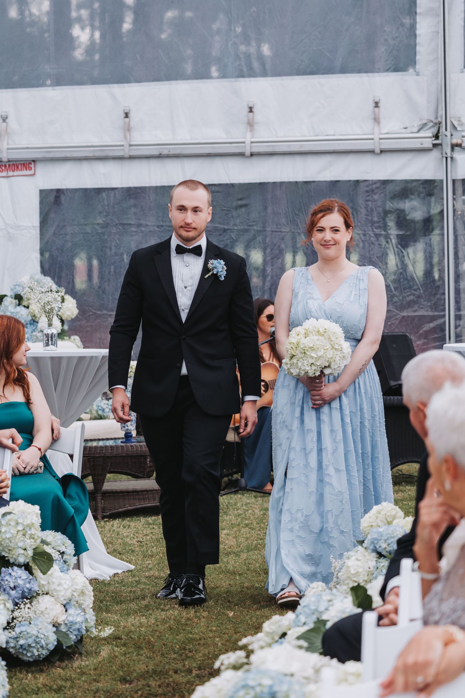 Groomsman escorts bridesmaid in light blue gown down the aisle at an elegant outdoor wedding ceremony.