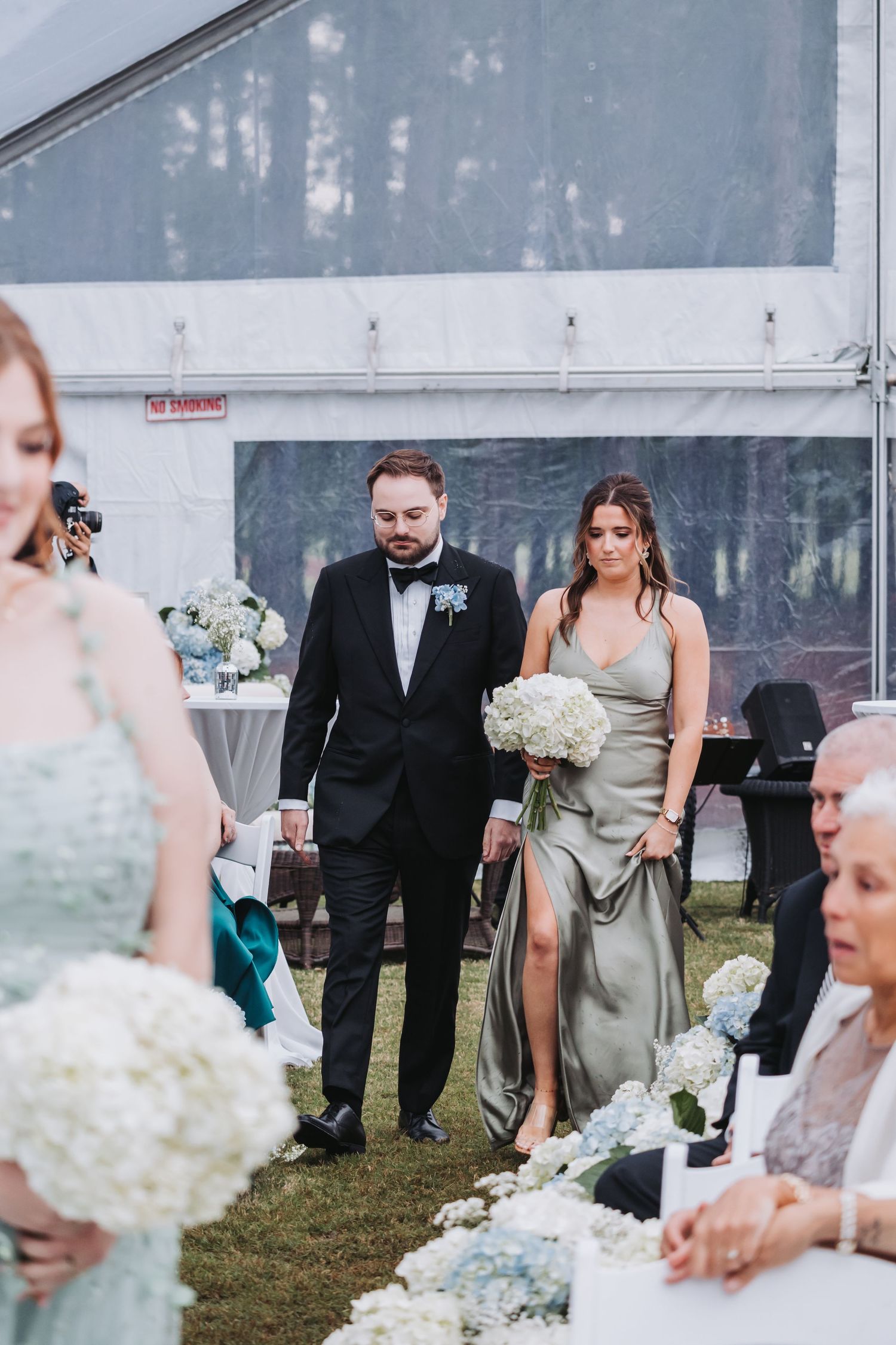 Groomsman escorting bridesmaid in sage green gown down aisle at outdoor tented wedding.