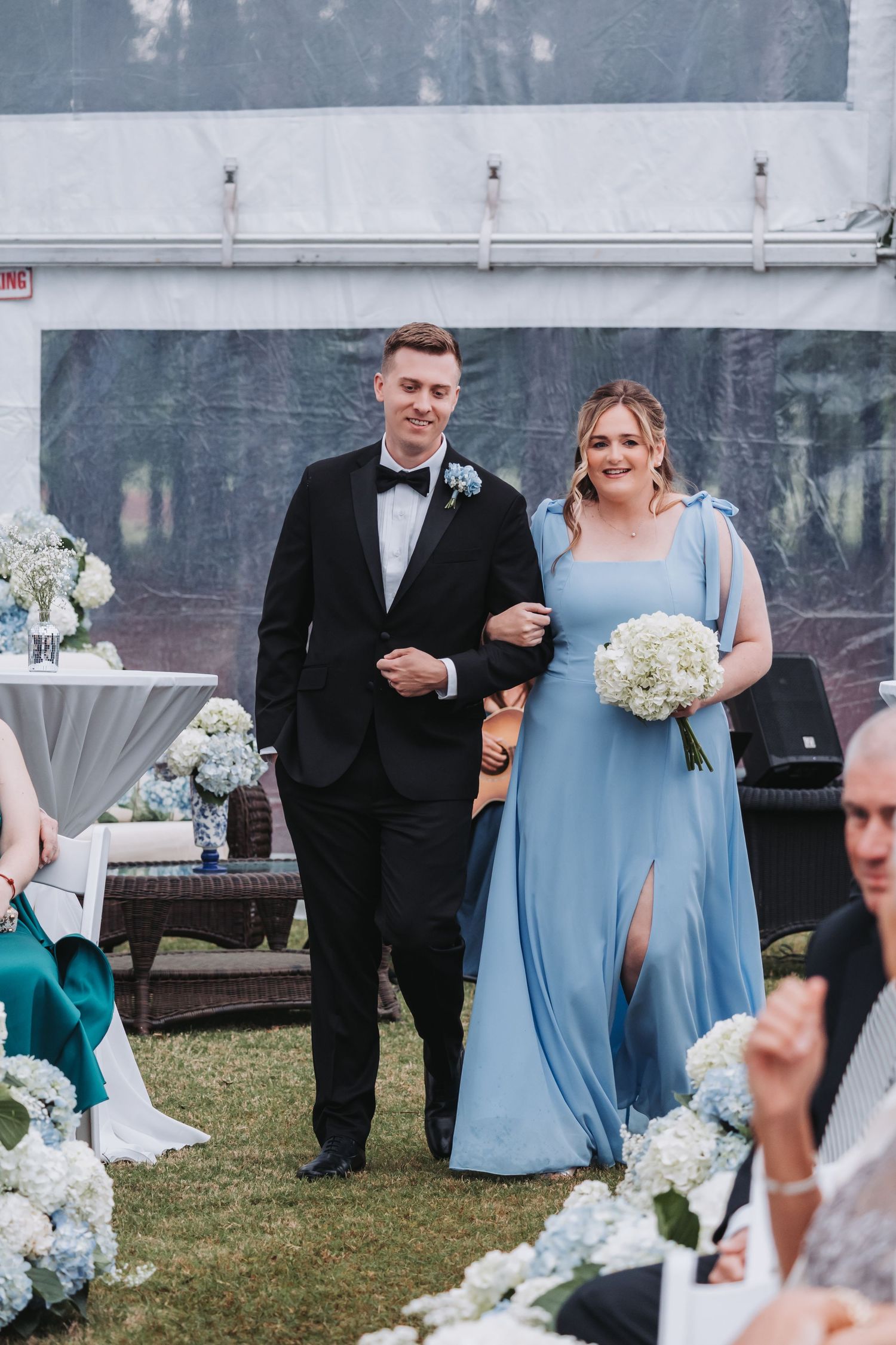 Groomsman in black tuxedo escorts bridesmaid in light blue gown down outdoor wedding aisle under white tent.
