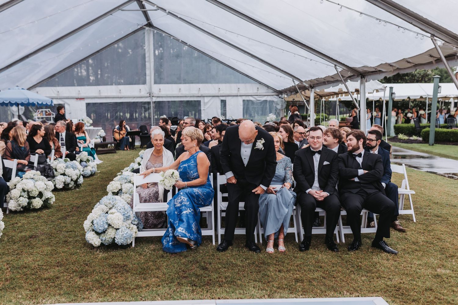 Wedding guests seated under a clear tent during an outdoor ceremony surrounded by white hydrangea floral arrangements.