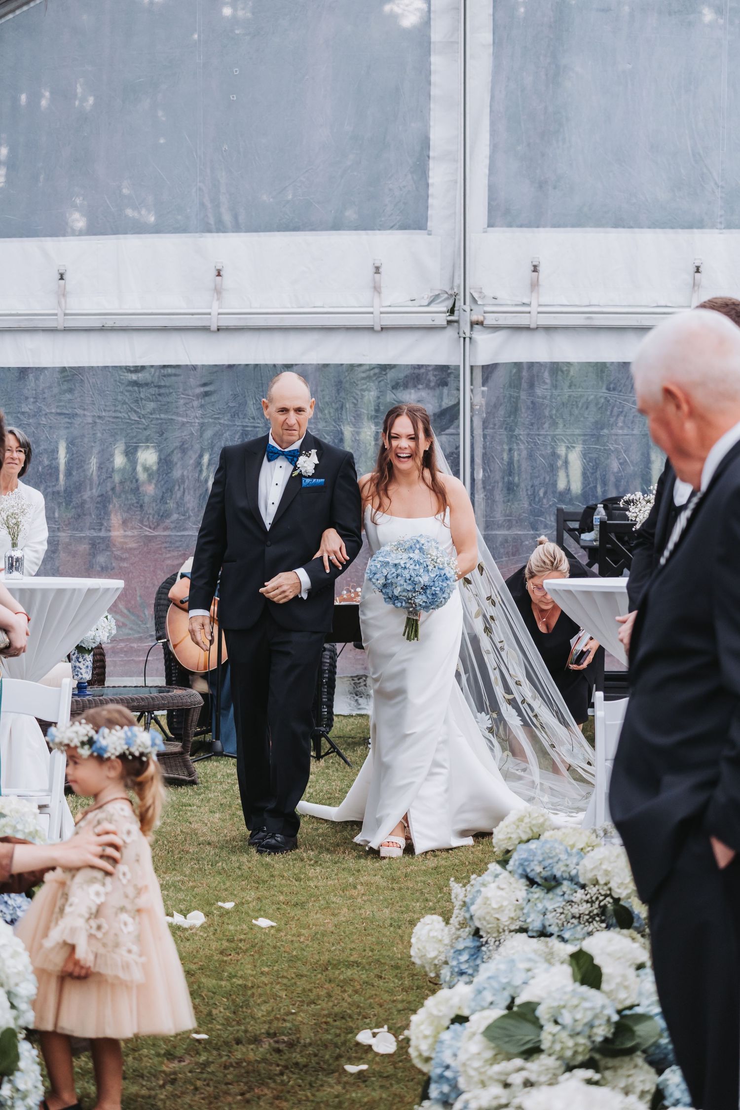 Bride in strapless white gown carrying blue hydrangea bouquet walks down aisle with escort at outdoor tented wedding.