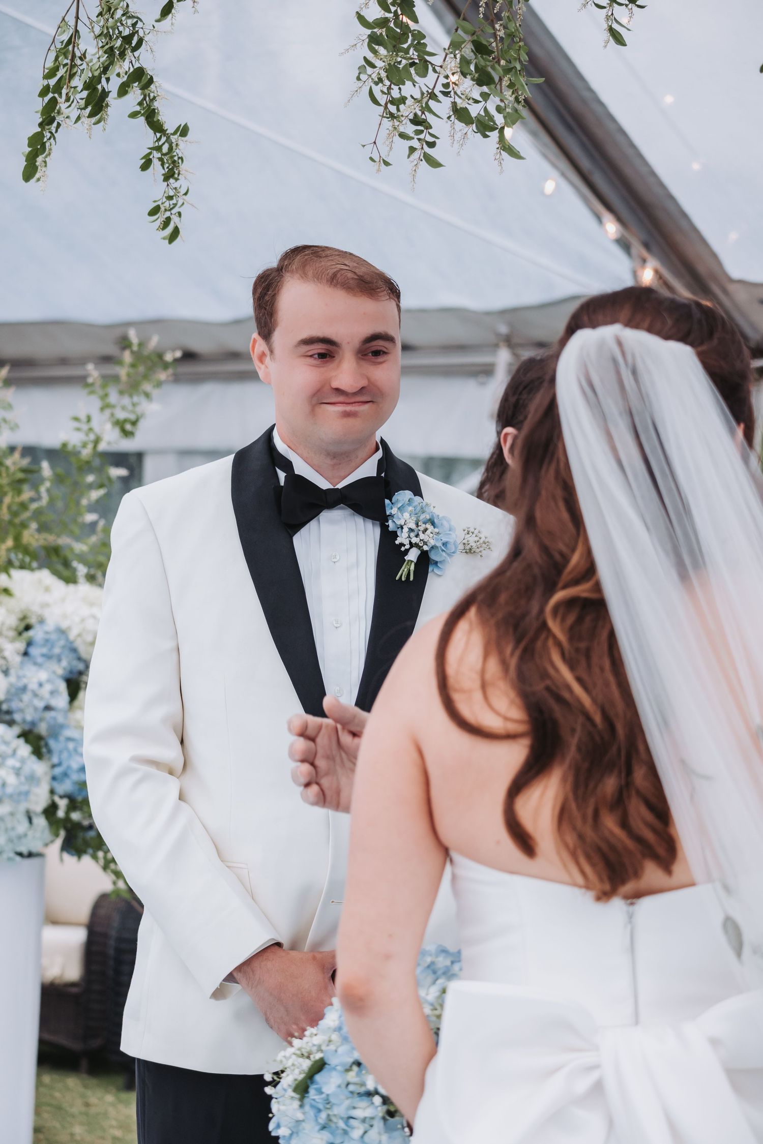 Groom in white tuxedo smiles at bride during outdoor wedding ceremony under a clear tent.