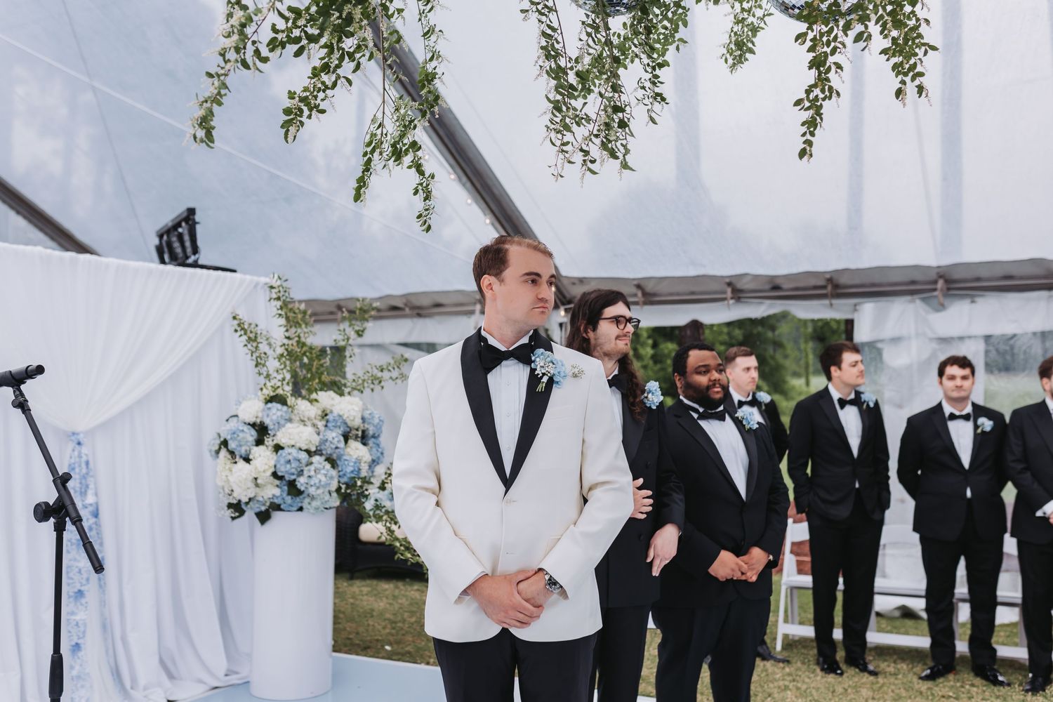 Groom in white tuxedo jacket stands at altar with groomsmen in black tuxedos at outdoor wedding ceremony.