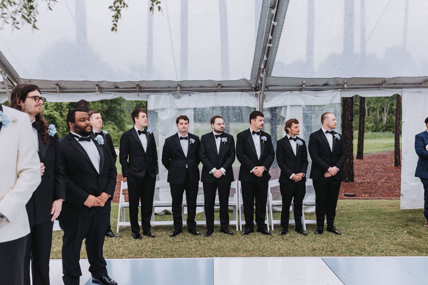 Groomsmen in black tuxedos with bow ties stand in a line at an outdoor wedding ceremony under a clear tent.
