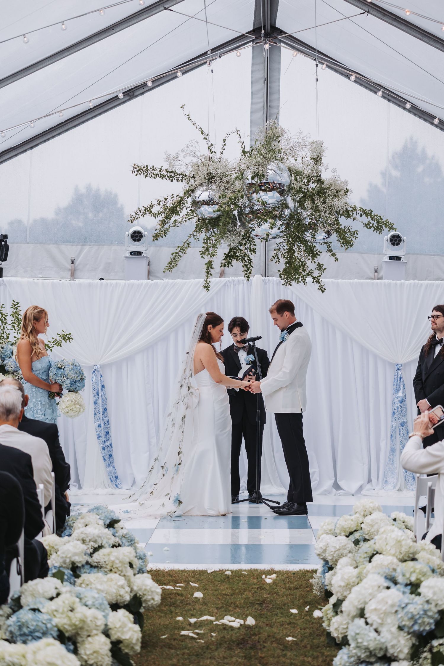 Bride and groom exchange vows at elegant outdoor wedding altar adorned with white draping and blue hydrangeas under clear tent.