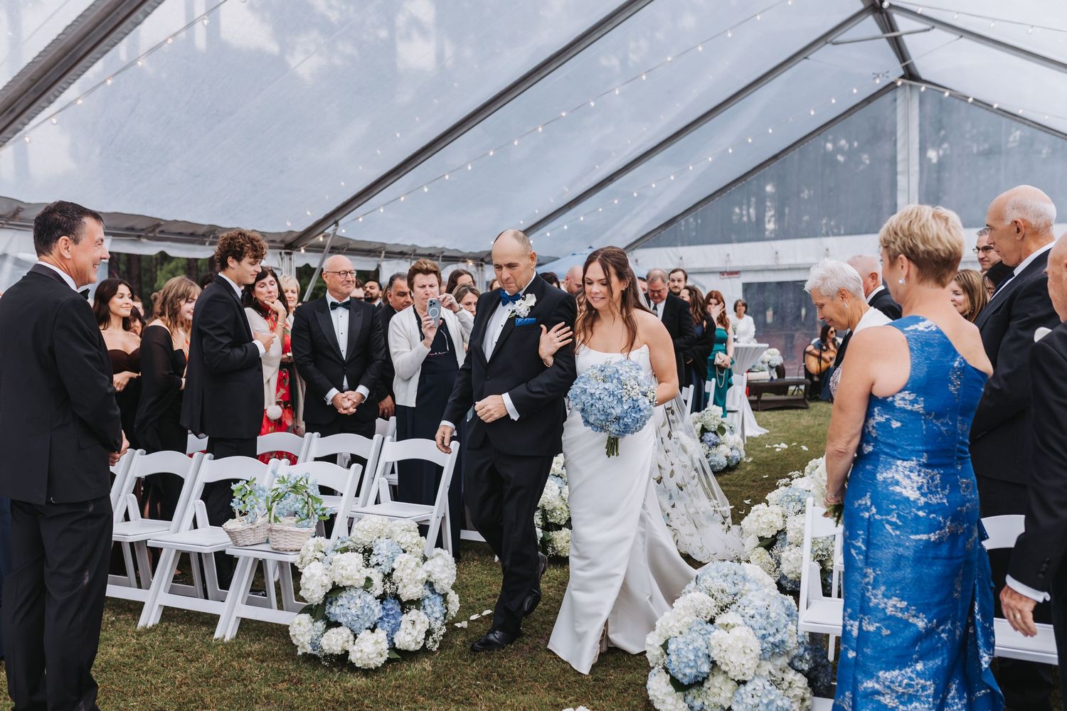 Bride walks down the aisle escorted by family under a clear wedding tent with hydrangea decor.