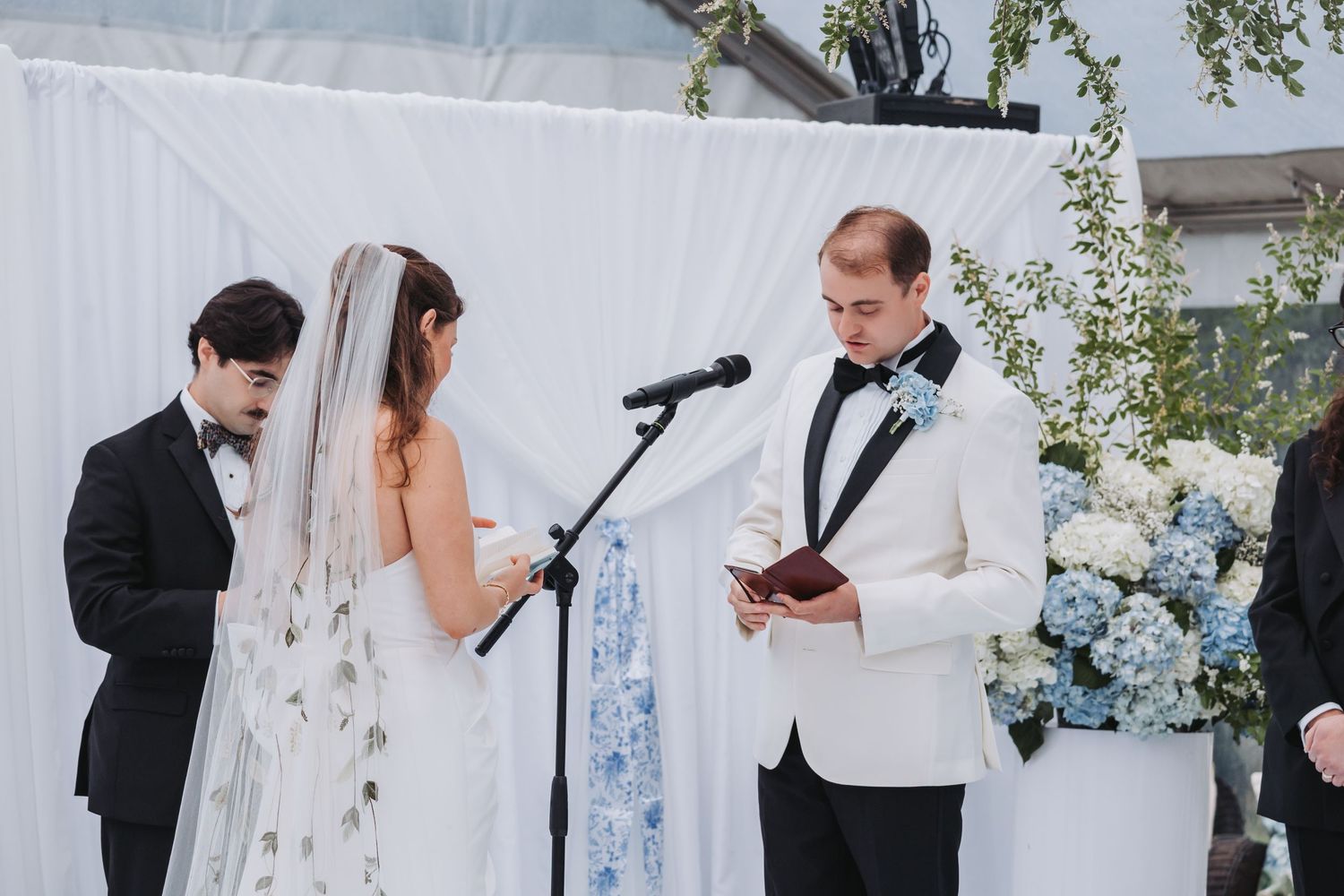 Groom in white tuxedo reading vows at outdoor wedding ceremony with floral backdrop.