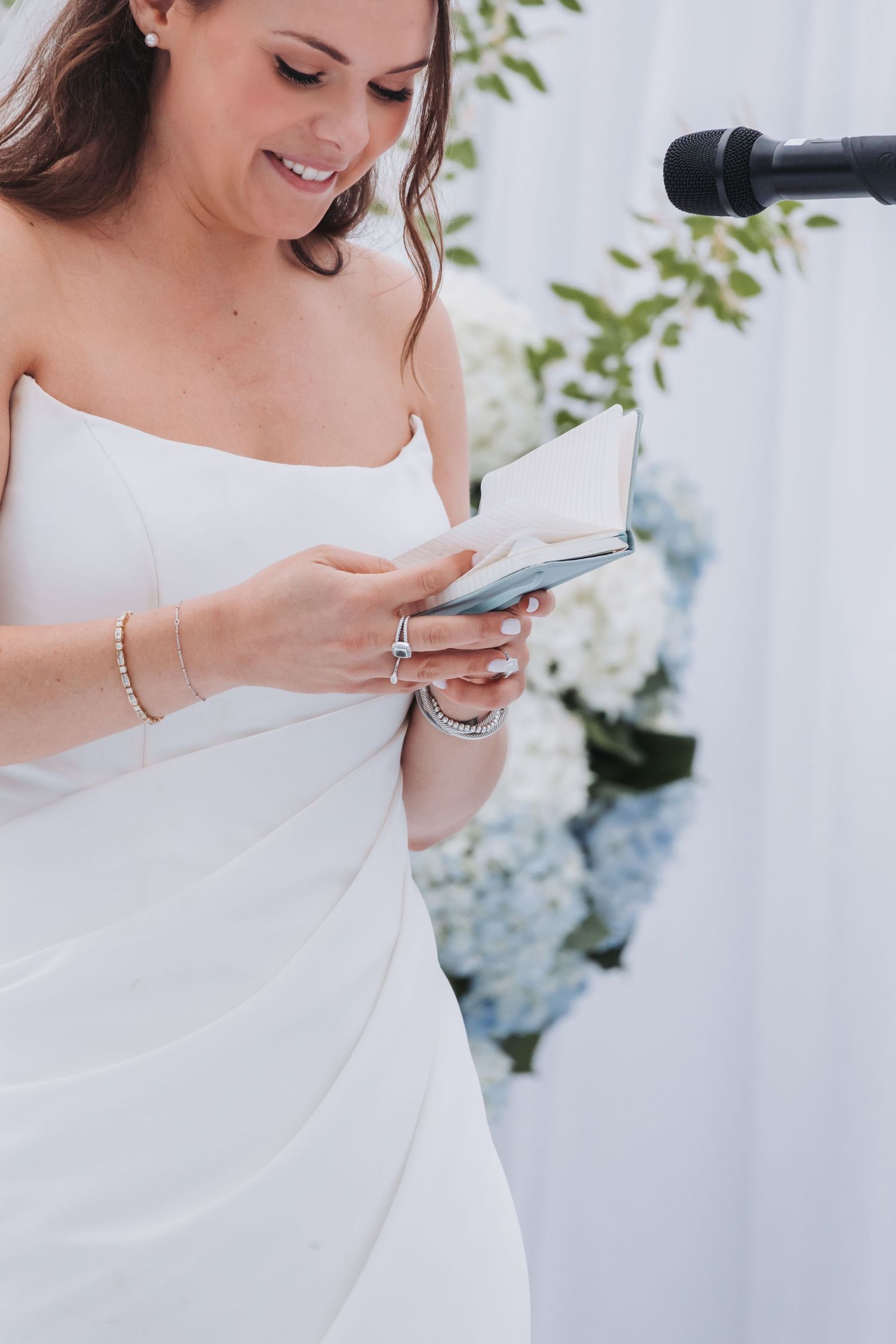 Smiling bride in strapless white gown reads personal vows from a small book at the wedding altar.