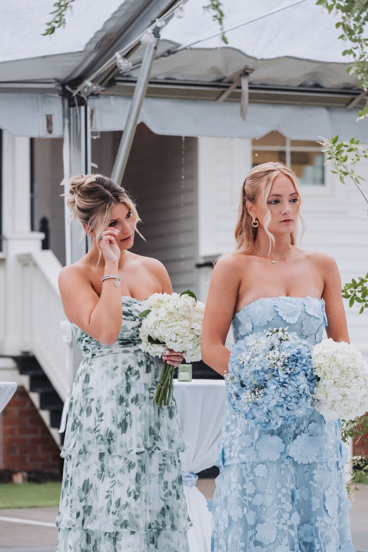 Two emotional bridesmaids in blue dresses holding white hydrangea bouquets during outdoor wedding ceremony.