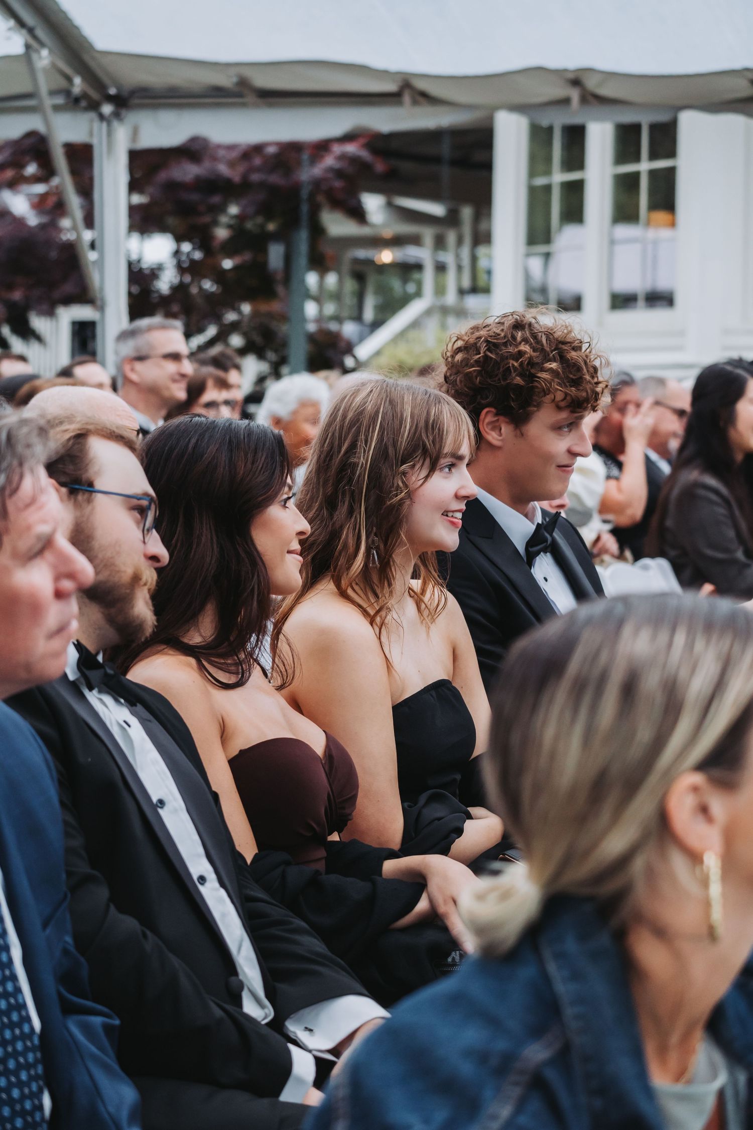 Smiling wedding guests in formal attire seated closely together during an elegant outdoor wedding ceremony.