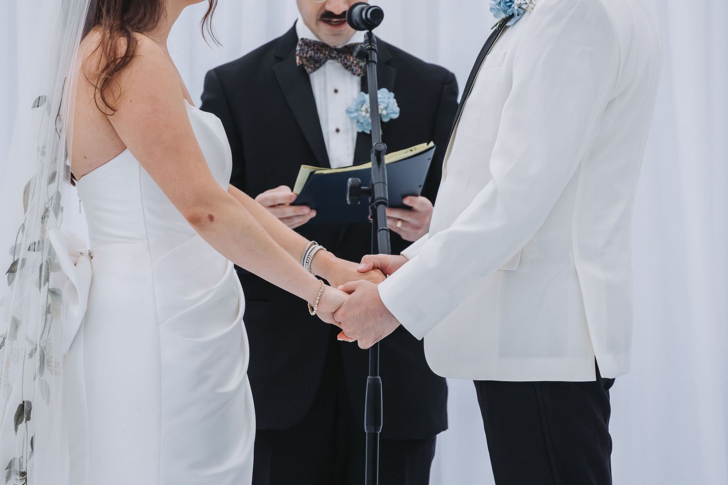 Couple holding hands at wedding altar with officiant in tuxedo and blue boutonniere during ceremony.