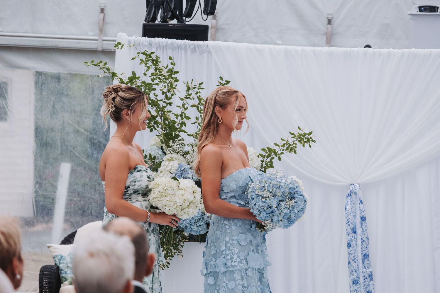 Two bridesmaids in light blue floral dresses holding white and blue hydrangea bouquets at an outdoor wedding ceremony.