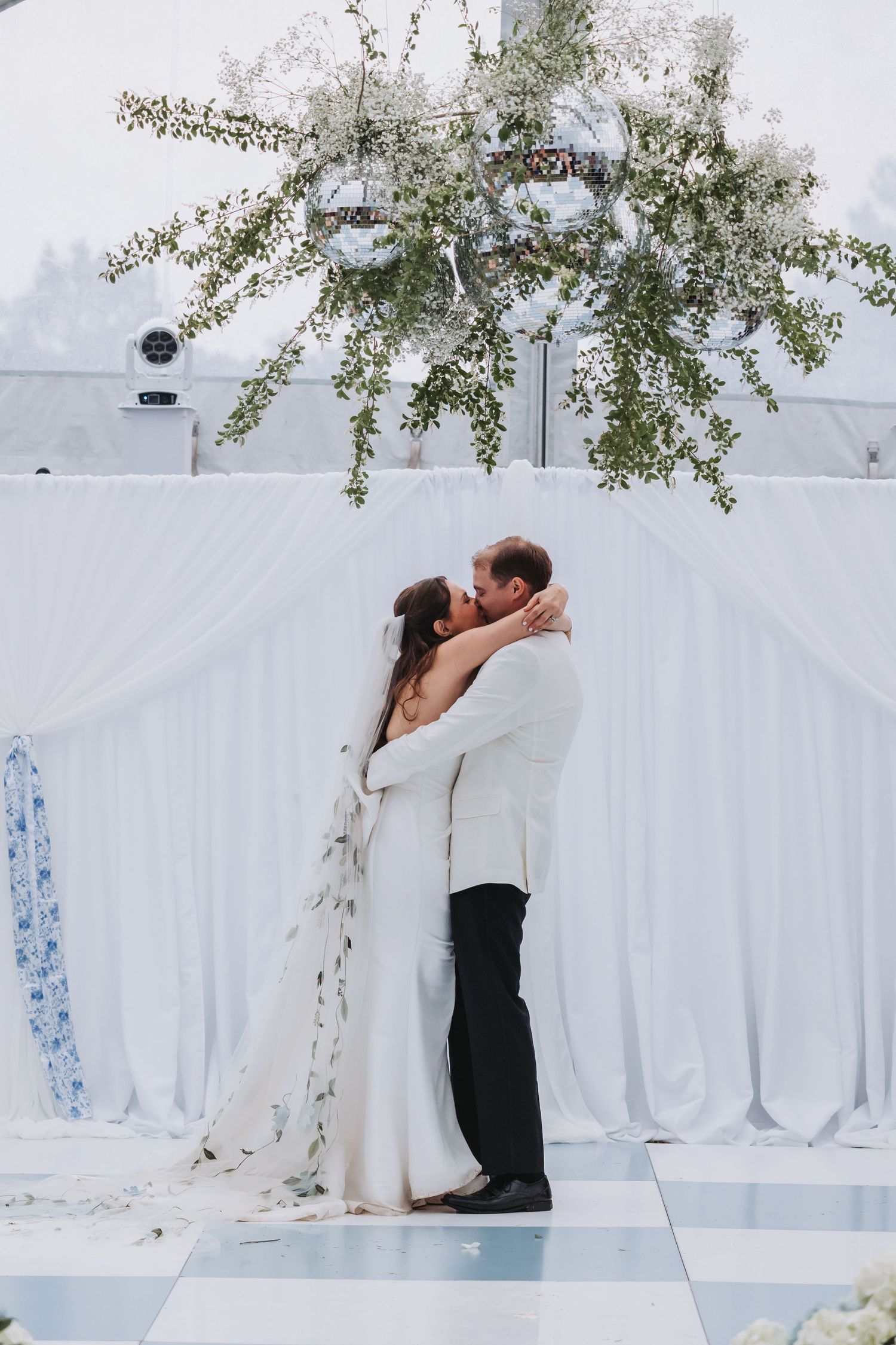 Bride and groom share first kiss under floral chandelier at elegant white wedding ceremony.