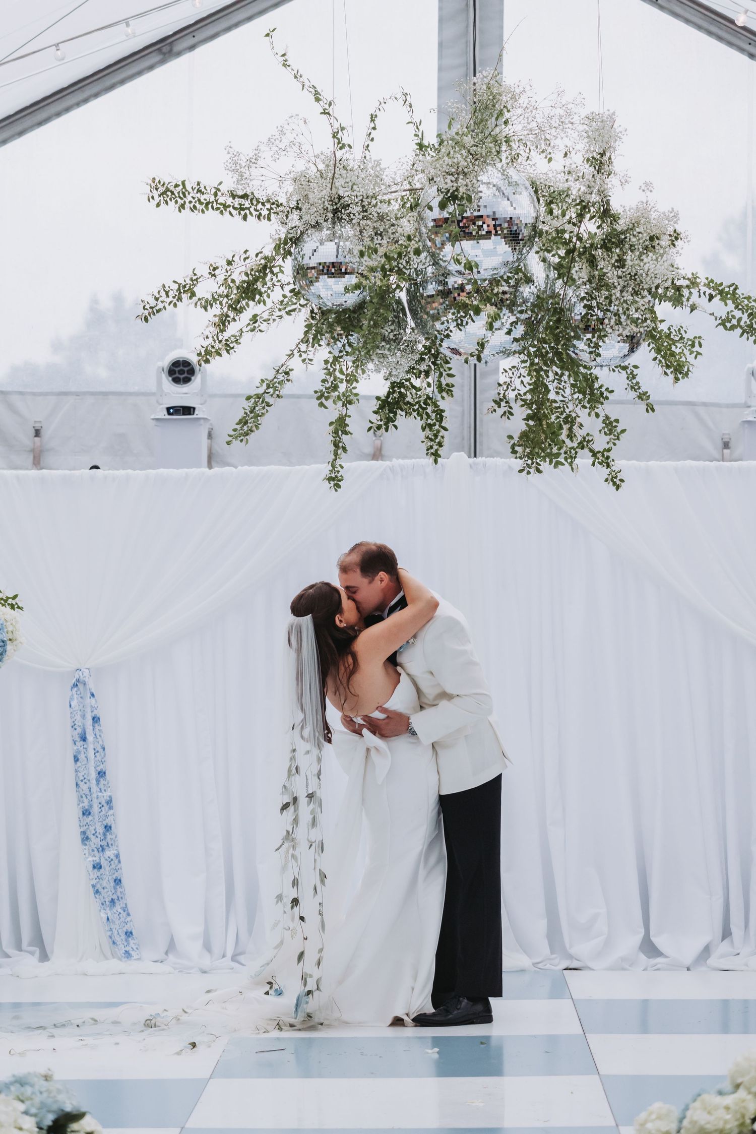 Bride and groom share a romantic first kiss under floral chandelier at elegant white wedding ceremony.