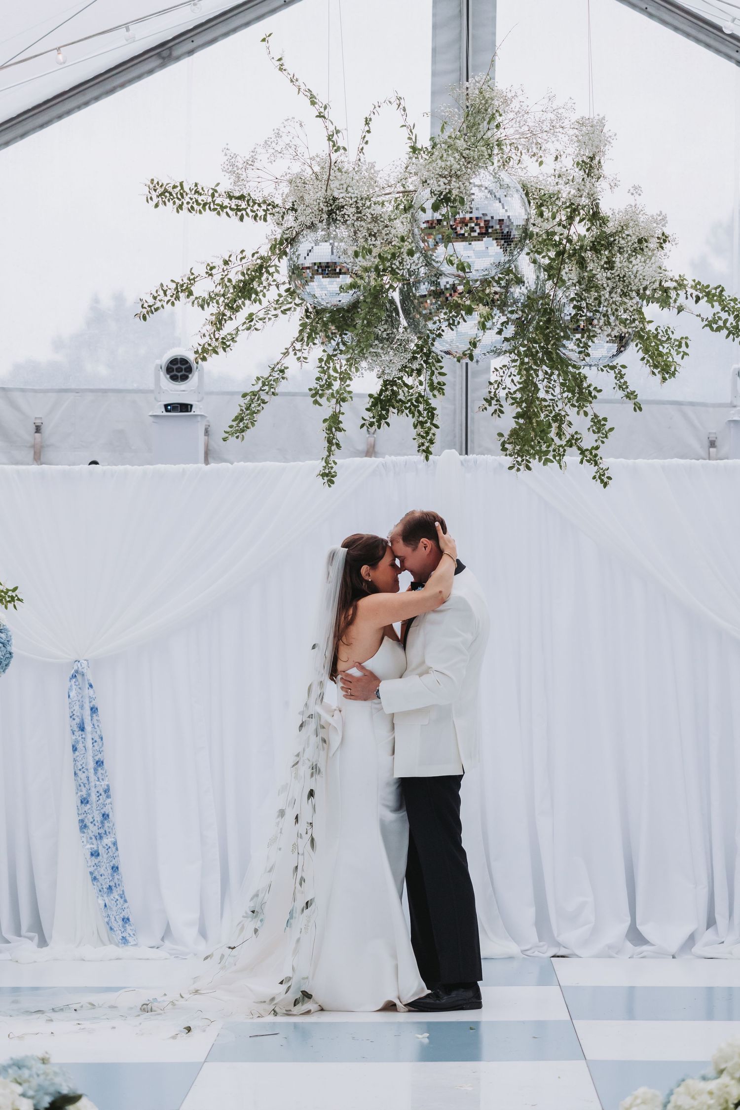 Bride and groom share romantic first dance under floral chandelier in elegant white draped wedding tent.