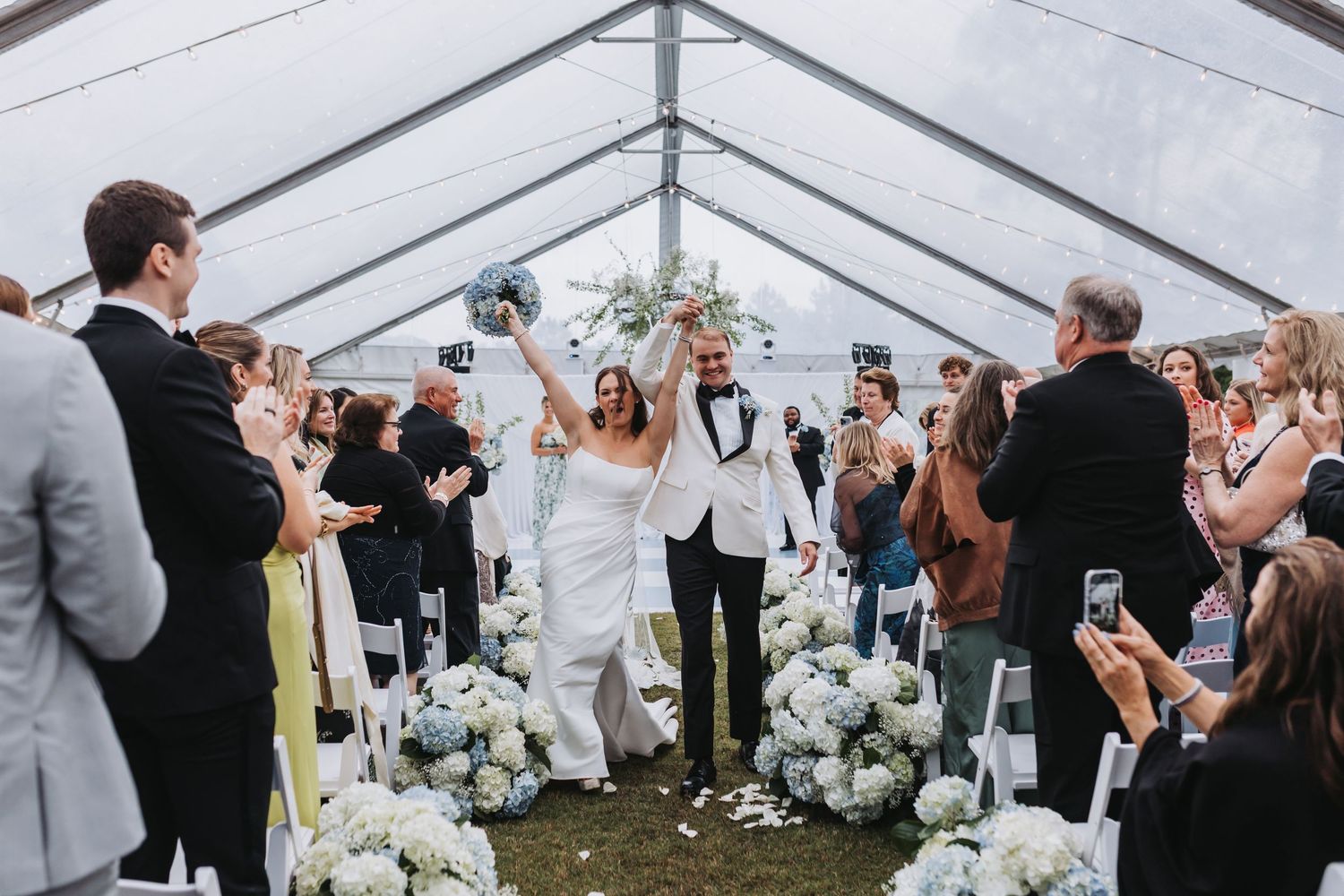 Joyful bride and groom walk down aisle under white tent, flanked by white floral arrangements and cheering guests.