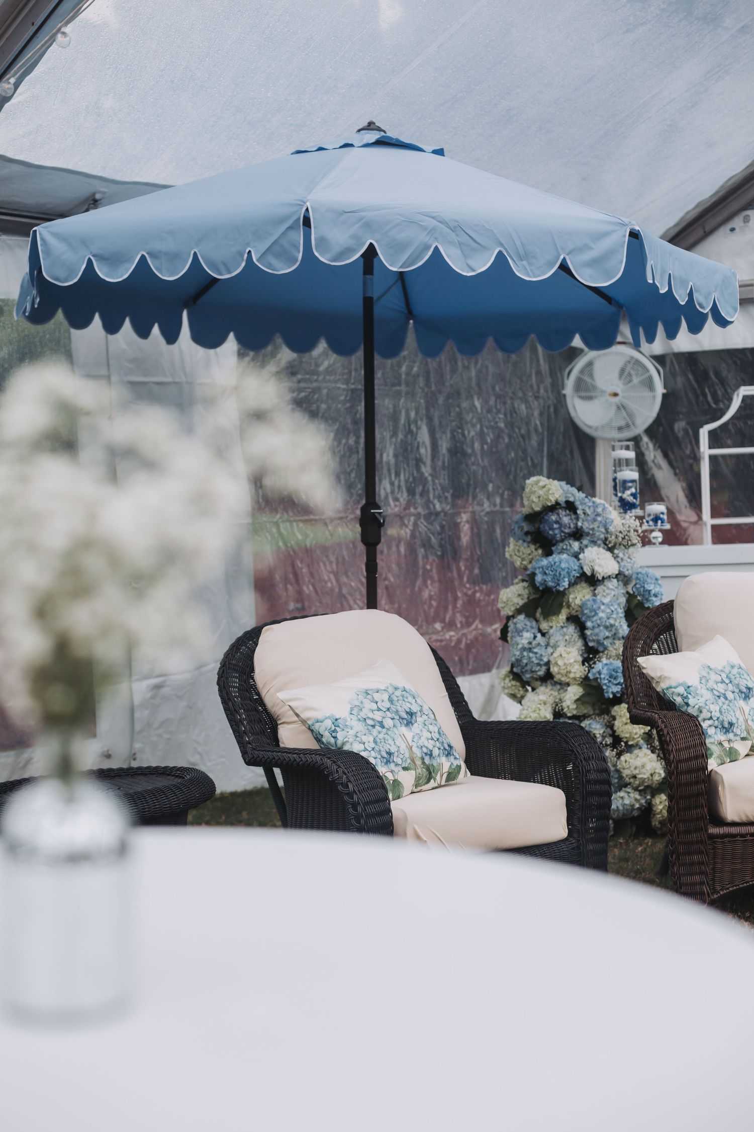 Blue scalloped patio umbrella over wicker chairs with floral cushions and hydrangea arrangements in a tent reception space.