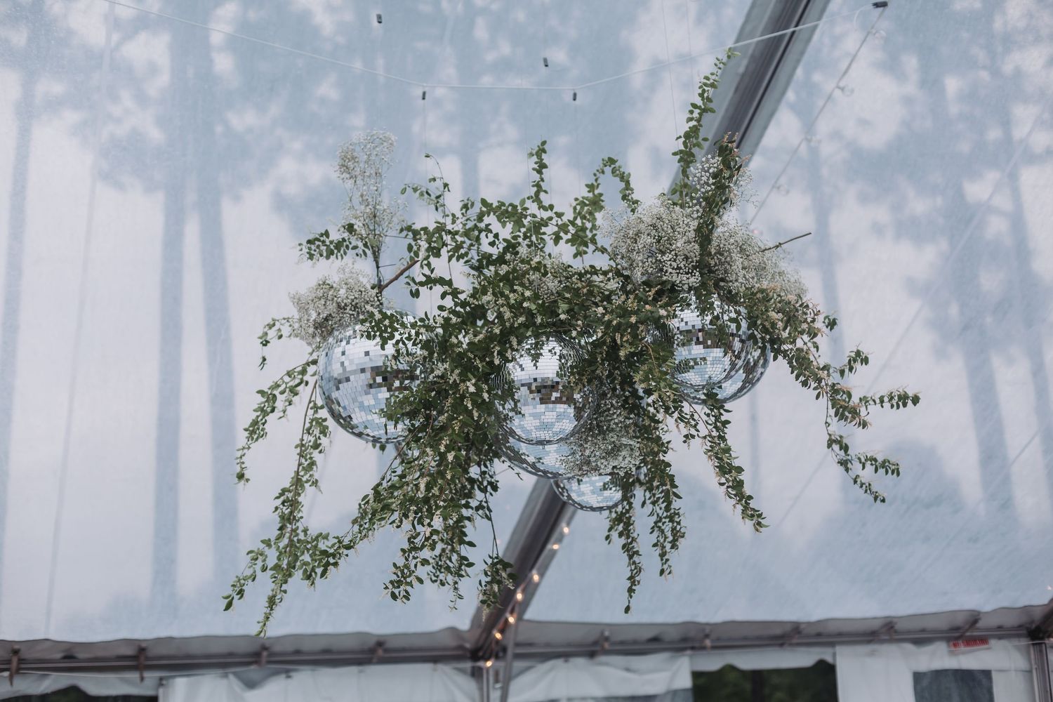 Lush greenery and white floral chandelier hanging from a clear wedding tent ceiling.