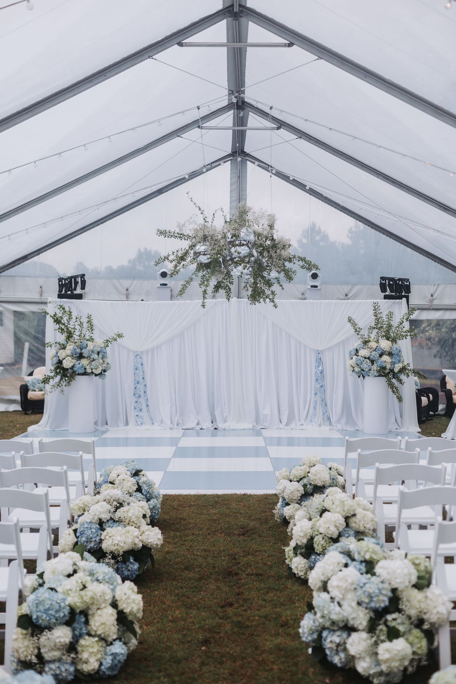 Elegant wedding ceremony tent with white draping, blue hydrangeas lining the aisle, and floral chandelier.