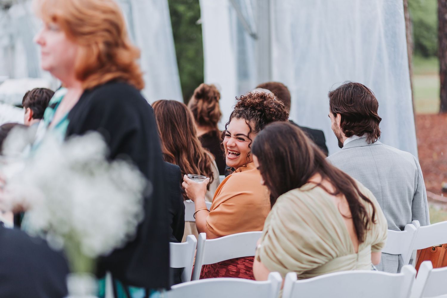 Smiling wedding guest hugs friend while seated in white chairs at outdoor tented wedding ceremony.