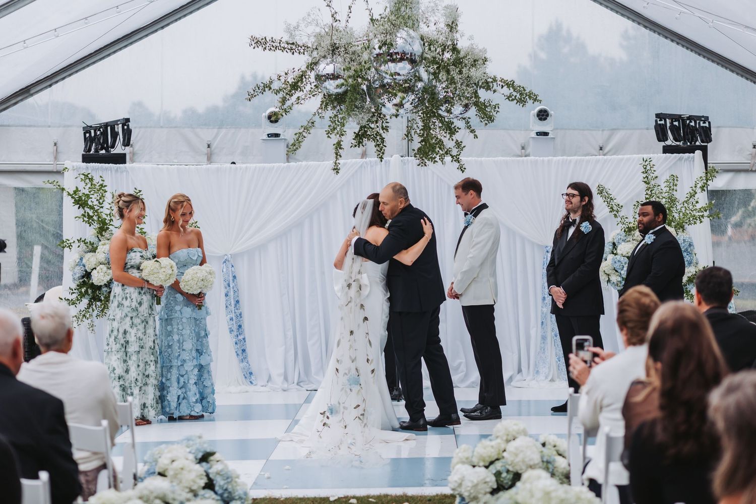 Bride and father share emotional hug at altar during tented outdoor wedding ceremony with blue florals.