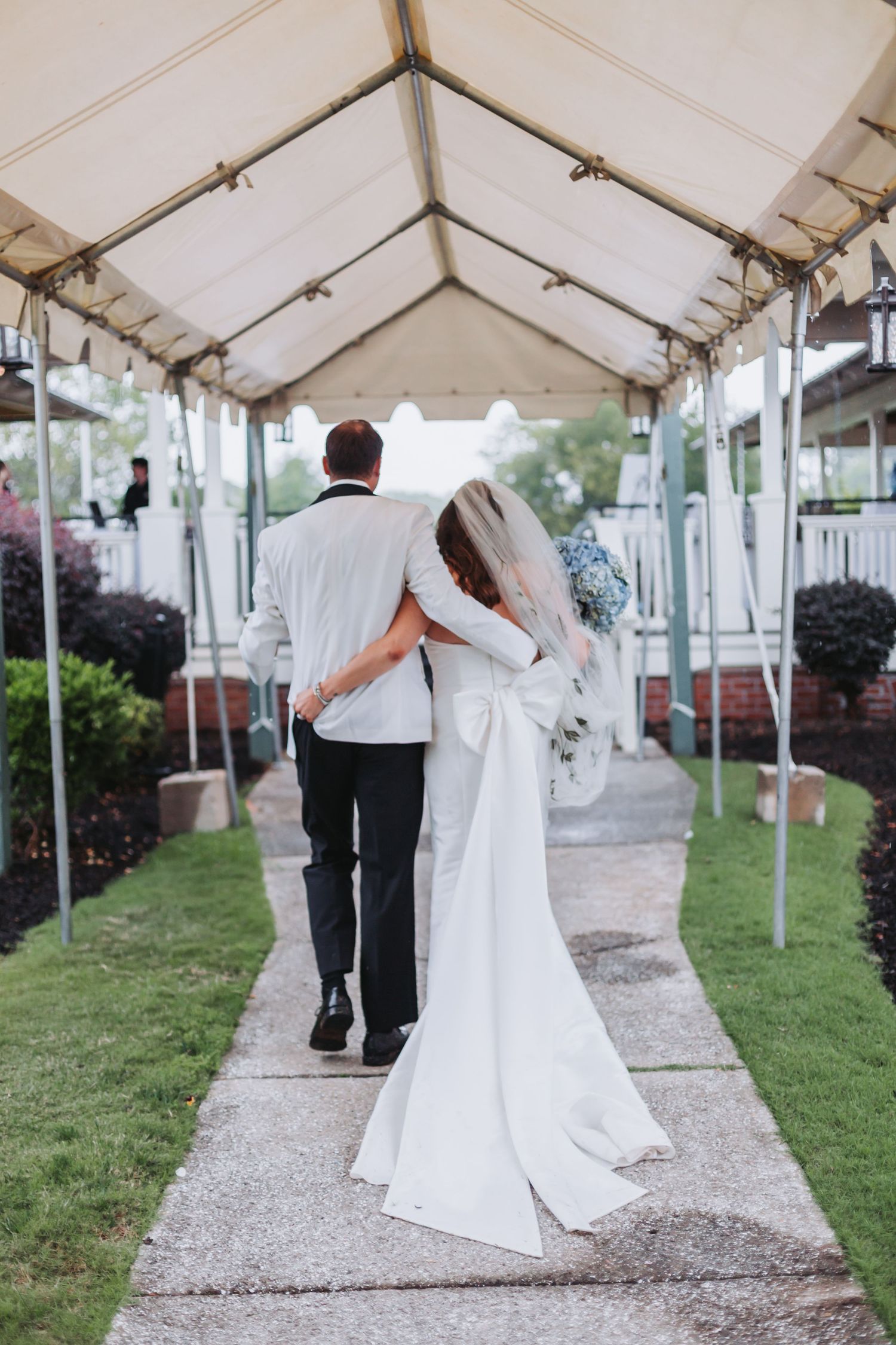Bride and groom walk arm in arm down a garden pathway under a white tent after ceremony.