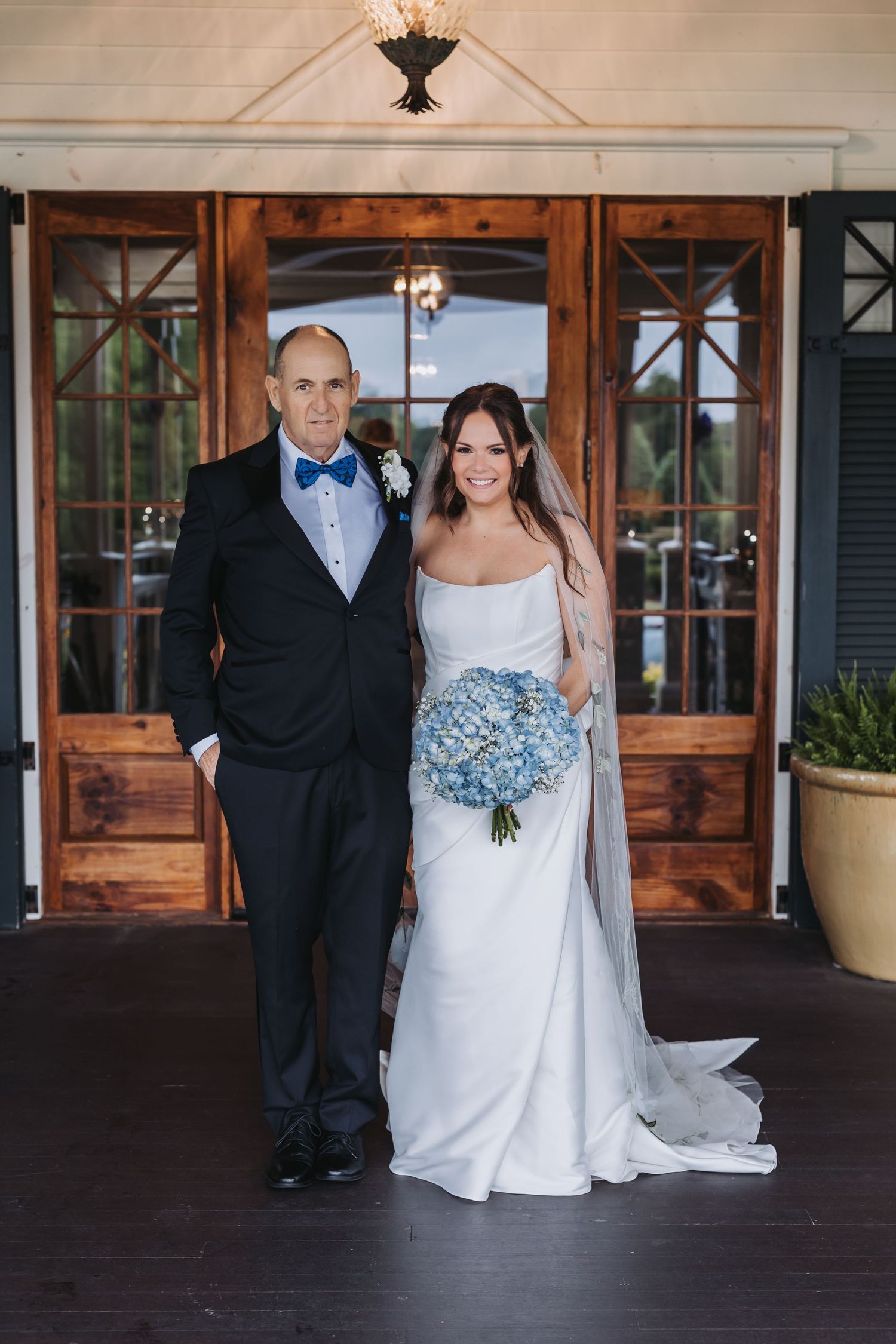 Bride in white gown holding blue hydrangea bouquet poses with father in black tuxedo before wooden doors.