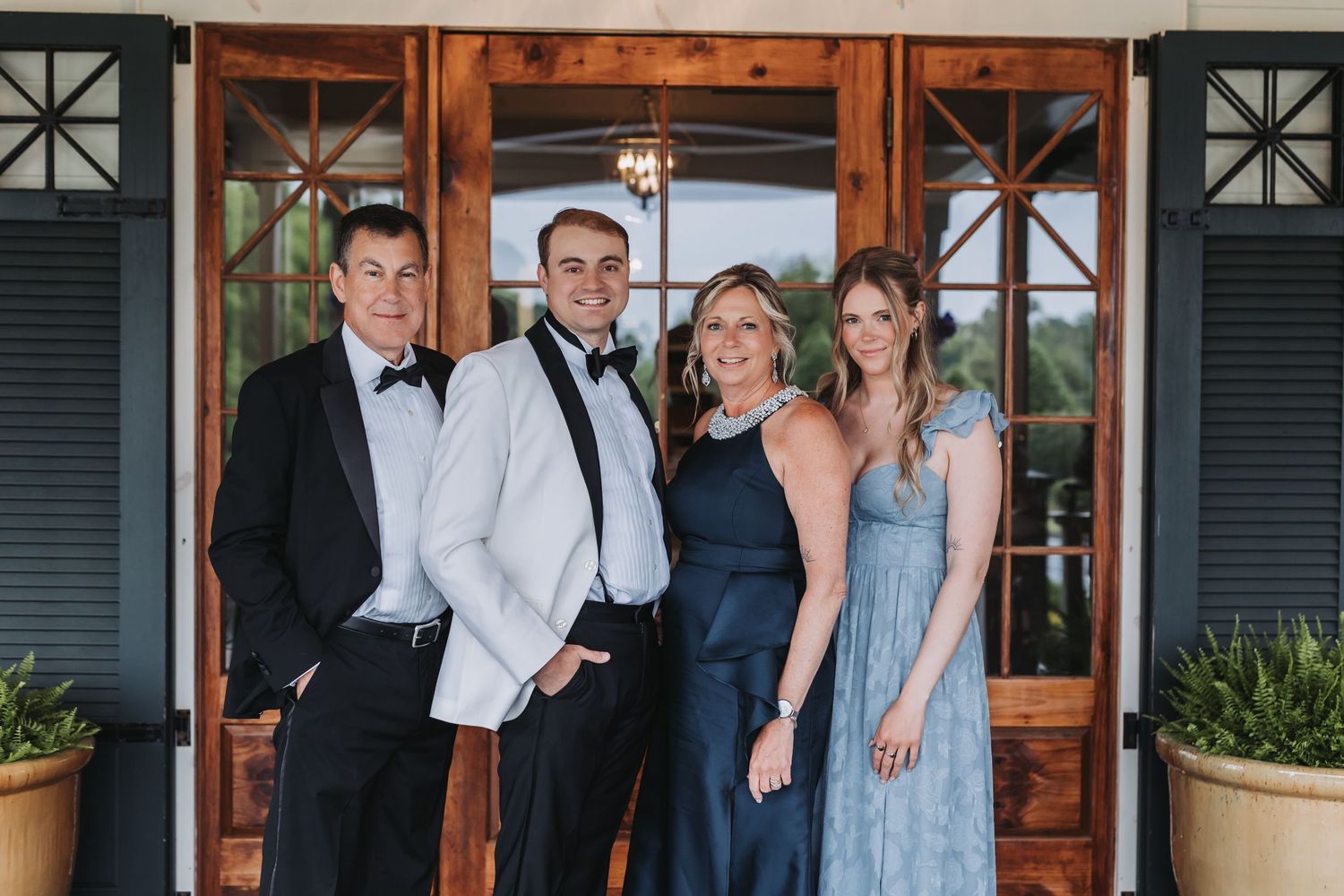 Family of four posing in formal attire before wooden doors at wedding venue.