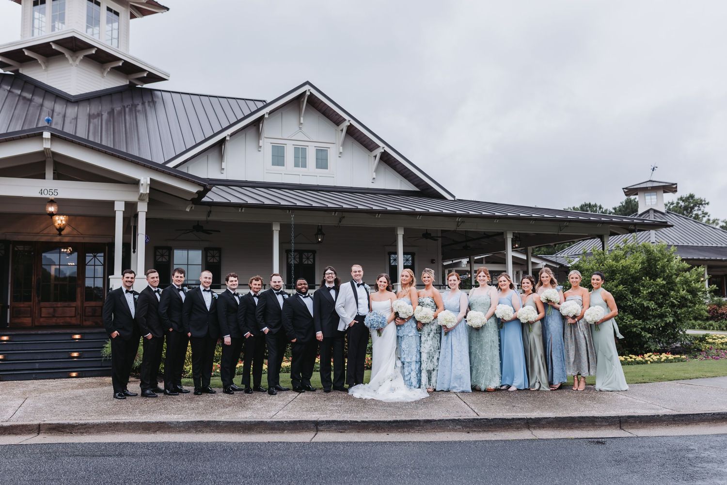 Large wedding party in black tuxedos and blue bridesmaid gowns poses outside venue.