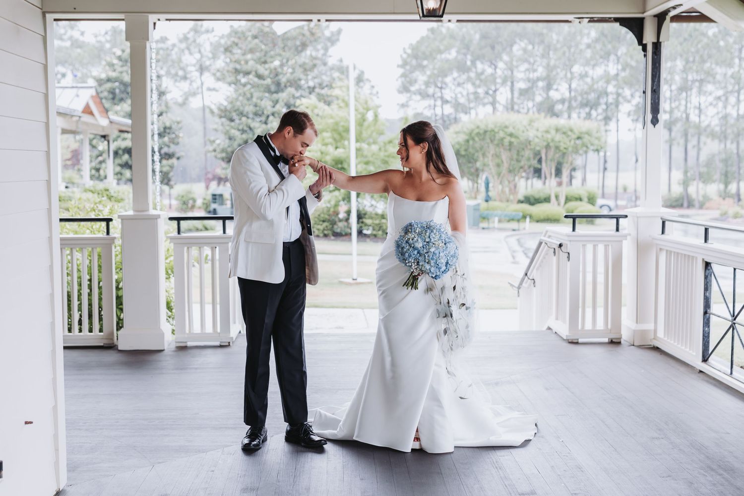Bride in strapless gown holding blue bouquet shares first look moment with groom on porch.
