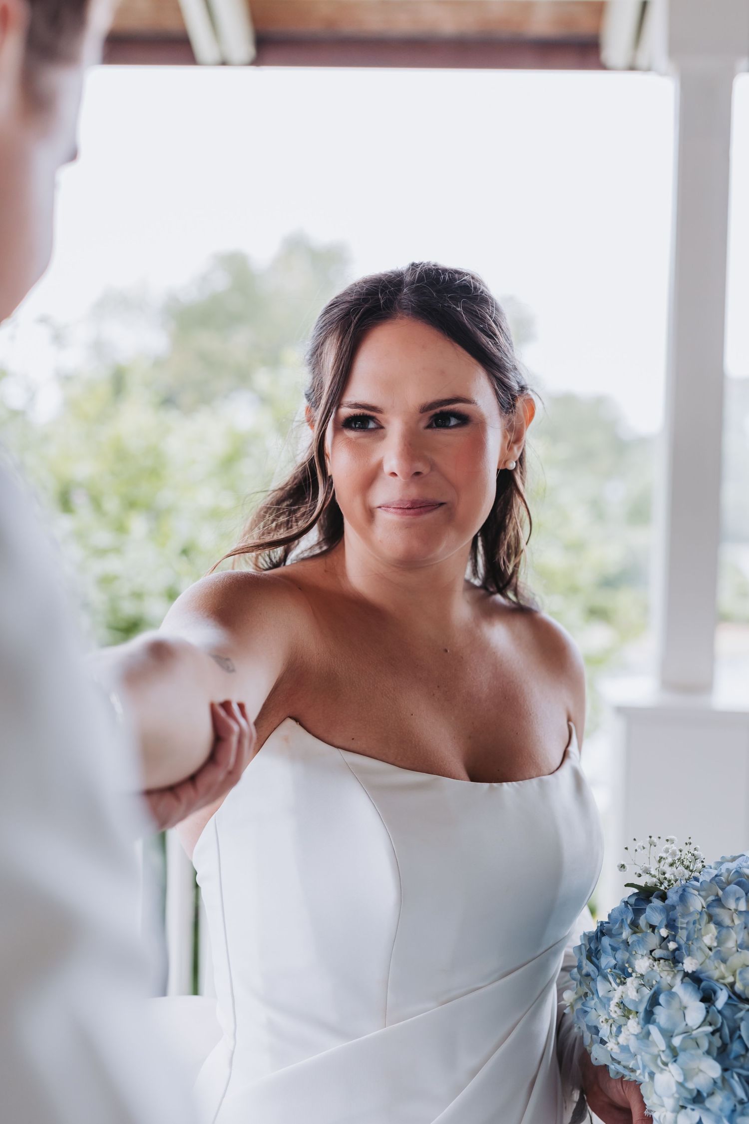 Radiant bride in strapless white gown holds blue hydrangea bouquet during outdoor wedding ceremony.