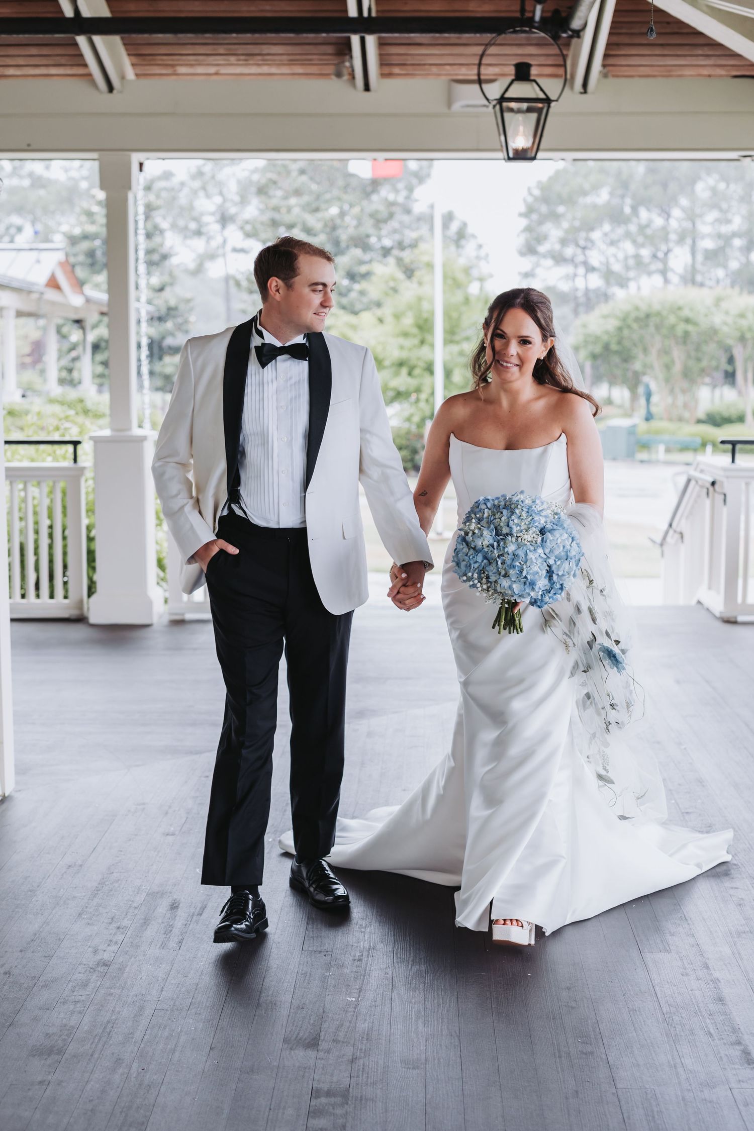 Bride in strapless white gown holding blue hydrangea bouquet walks hand-in-hand with groom in white tuxedo jacket.