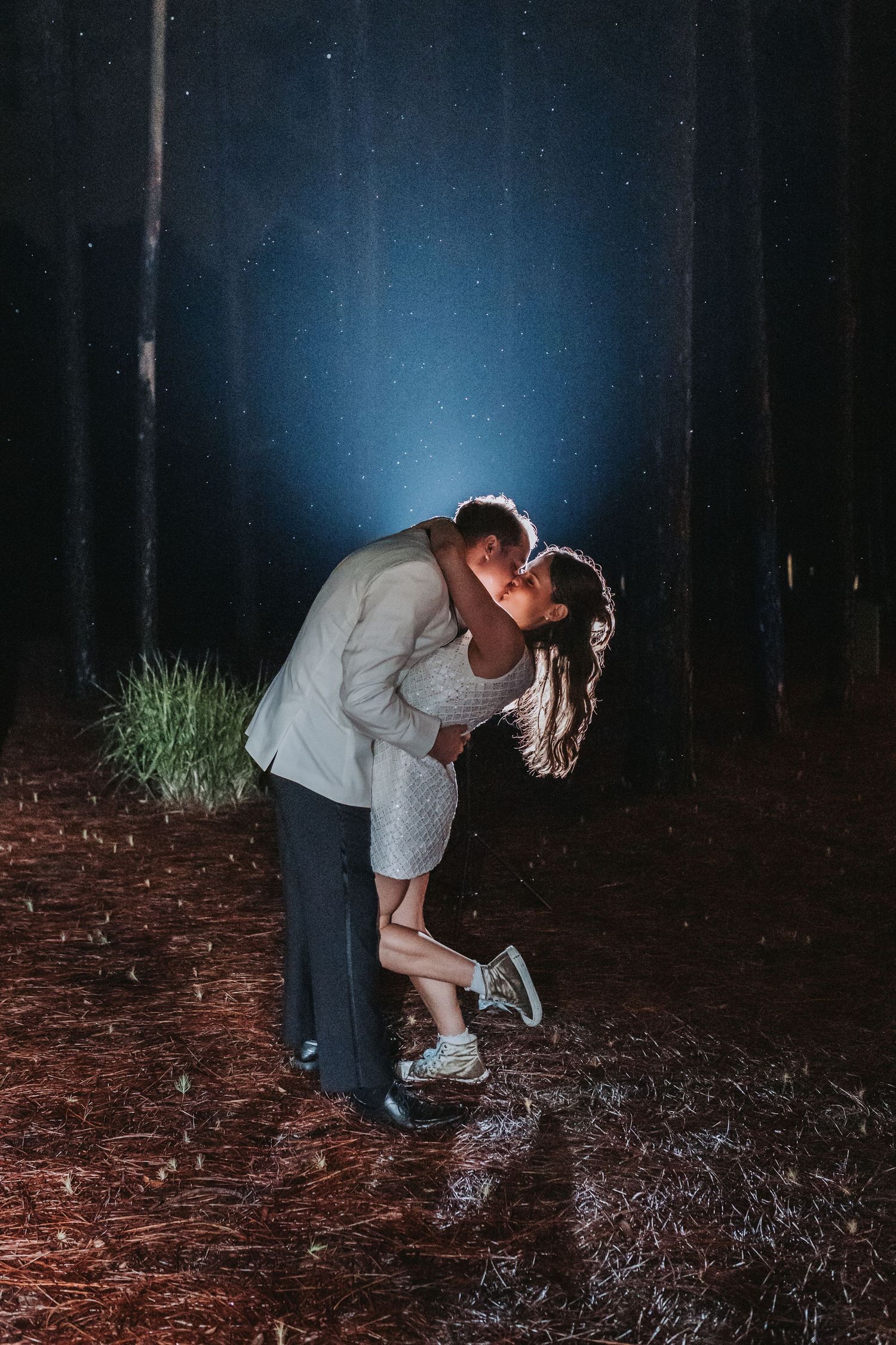 Groom dips bride for a romantic kiss in a dark forest illuminated by a single spotlight at night.