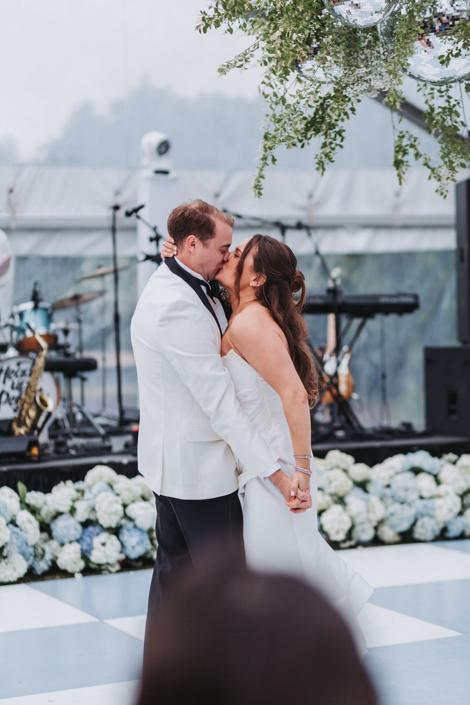 Bride and groom share first kiss on checkered dance floor at outdoor wedding reception with live band stage.