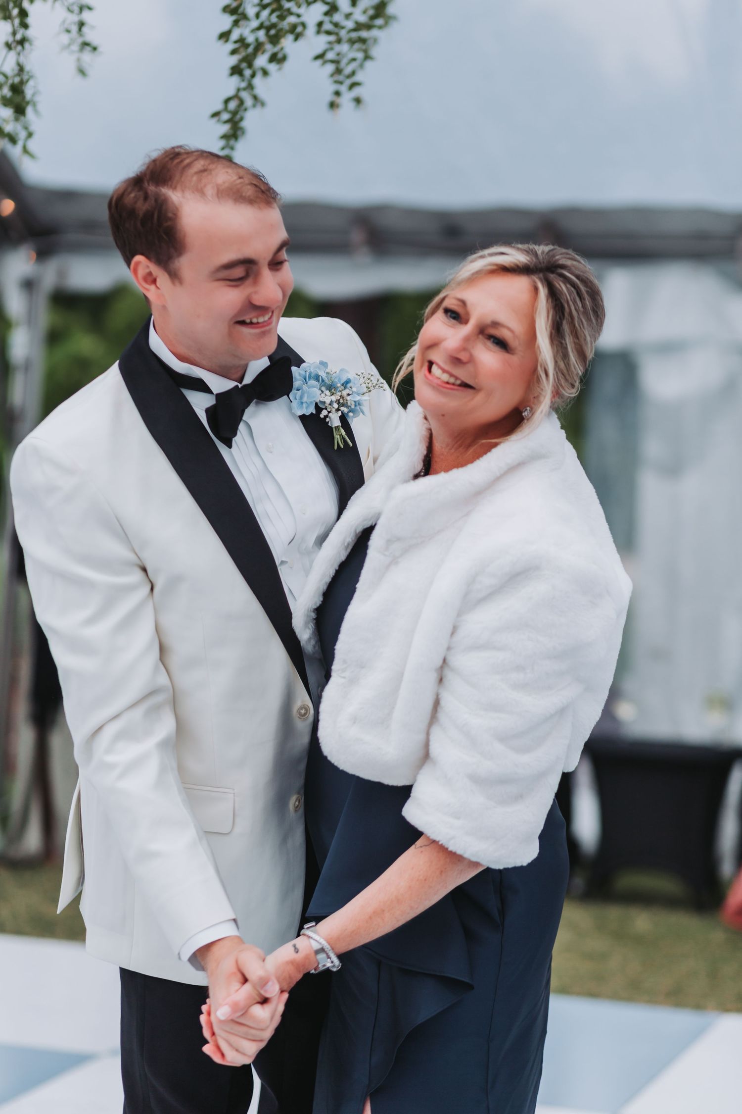 Smiling groom in white tuxedo posing with his mother in a white fur wrap outdoors.