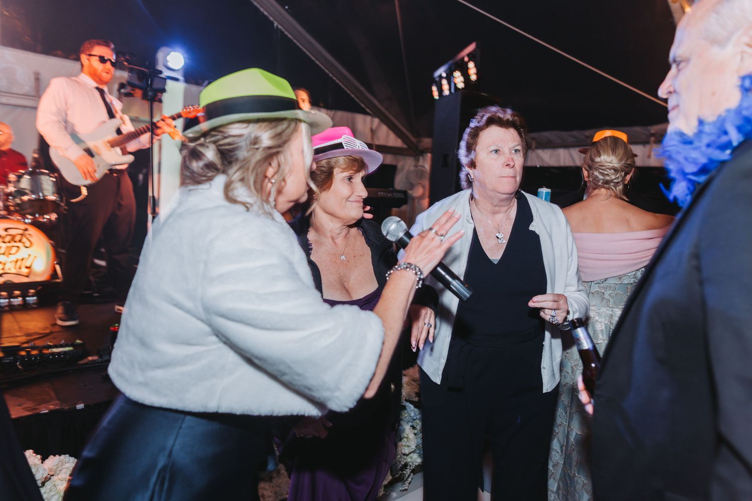 Wedding reception guests dance and sing with microphone while wearing colorful hats at lively evening party.