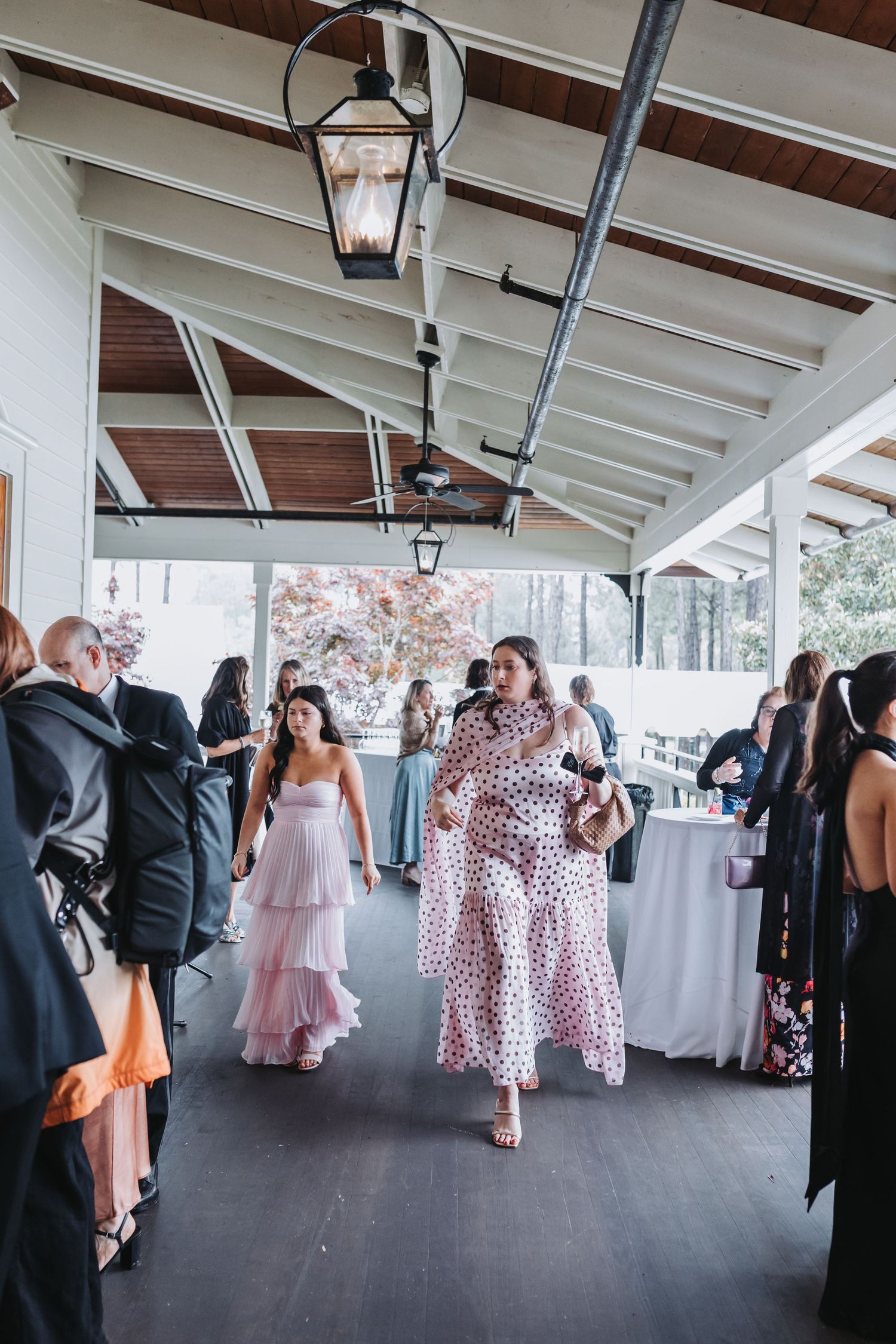 Wedding guests in formal attire mingle under a covered outdoor pavilion with hanging lanterns.