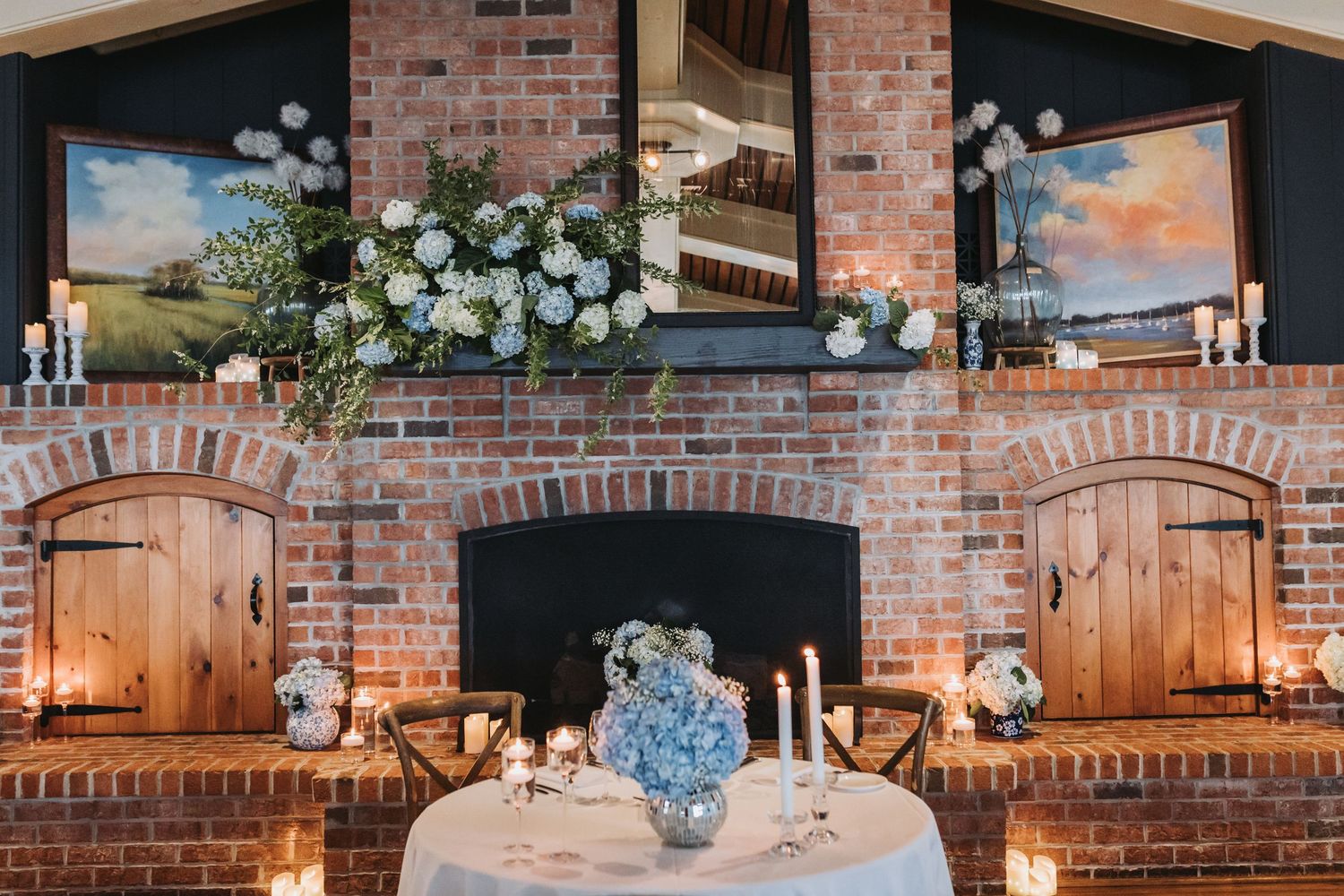 Elegant wedding sweetheart table set before a brick fireplace adorned with white floral arrangements and candles.