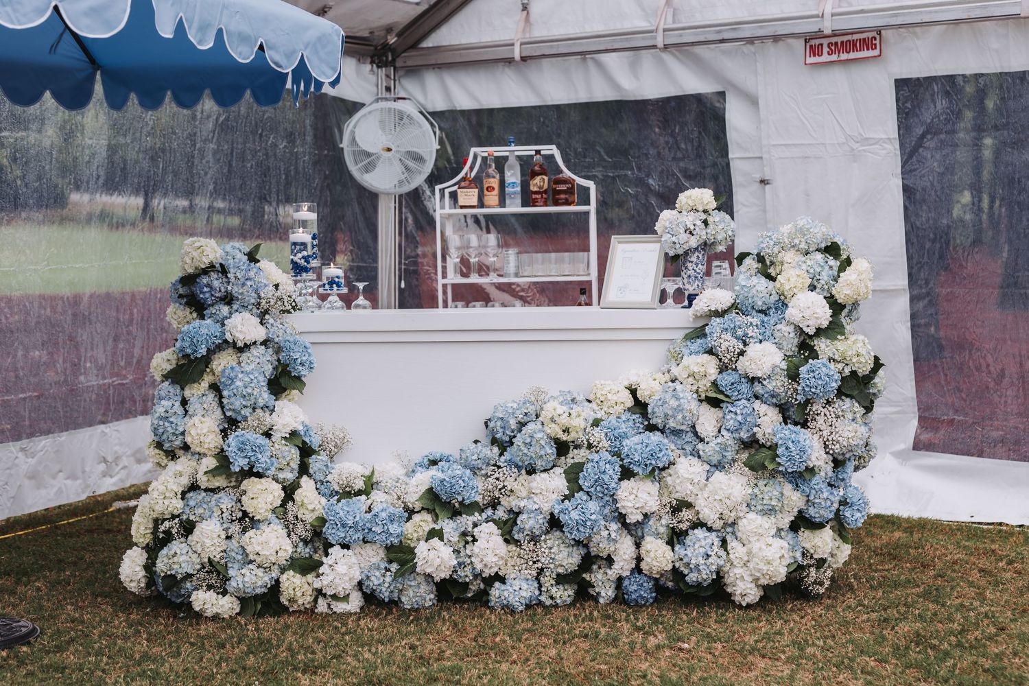 An outdoor wedding bar decorated with lush blue and white hydrangea floral arrangements under a tent.